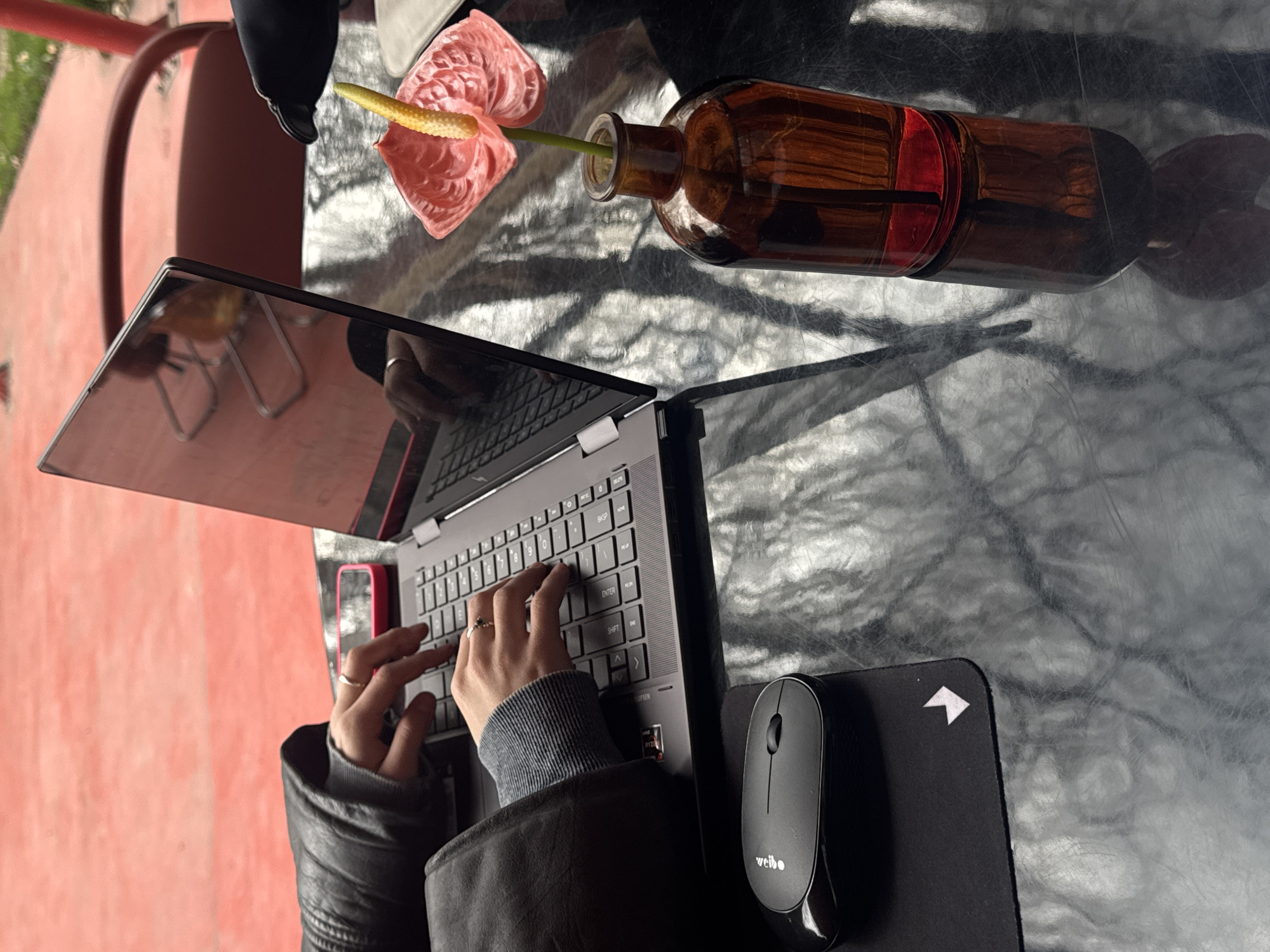 The image depicts an outdoor scene in Yerevan, Armenia, focusing on a workstation setup on a reflective, dark-colored table. A person, whose hands are visible, is actively typing on a dark grey laptop with a backlit keyboard. The individual wears a dark leather jacket with ribbed grey cuffs and silver rings on both ring fingers. To the left of the laptop, a black wireless mouse rests on a black mousepad featuring a white arrow icon in its lower right corner. A pink smartphone is positioned behind the laptop. To the right of the laptop, a dark amber glass vase contains a single pink Anthurium flower with a prominent yellow spadix, and a small amount of red liquid is visible at the vase's base. The table's surface reflects the sky, distant structures, and foliage. The background features a reddish-orange ground or paved area, with a partial dark red chair frame and a hint of green grass visible in the upper right.