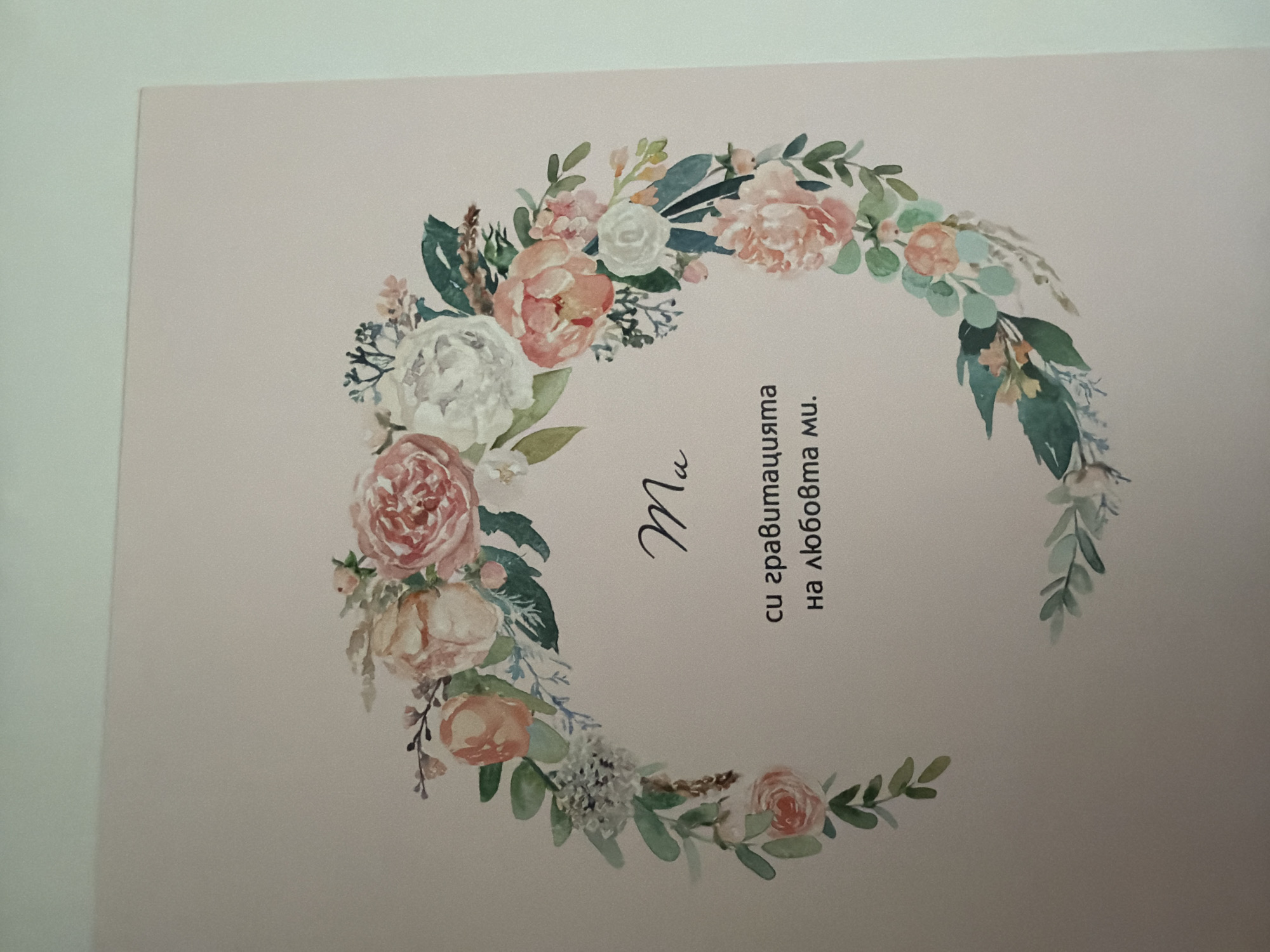 The image shows a close-up of a card with a floral wreath design. The text on the card, written in what appears to be Cyrillic script, is centered and appears to read "Ma" on the first line, followed by "си гравитацията" on the second line, and "на любовта ми." on the third line. The background is a pale pink color. The floral wreath is composed of watercolor illustrations of pink and white roses, green leaves, and other small flowers. The image does not contain any information about the city of Pazardzhik, Bulgaria, or any people.