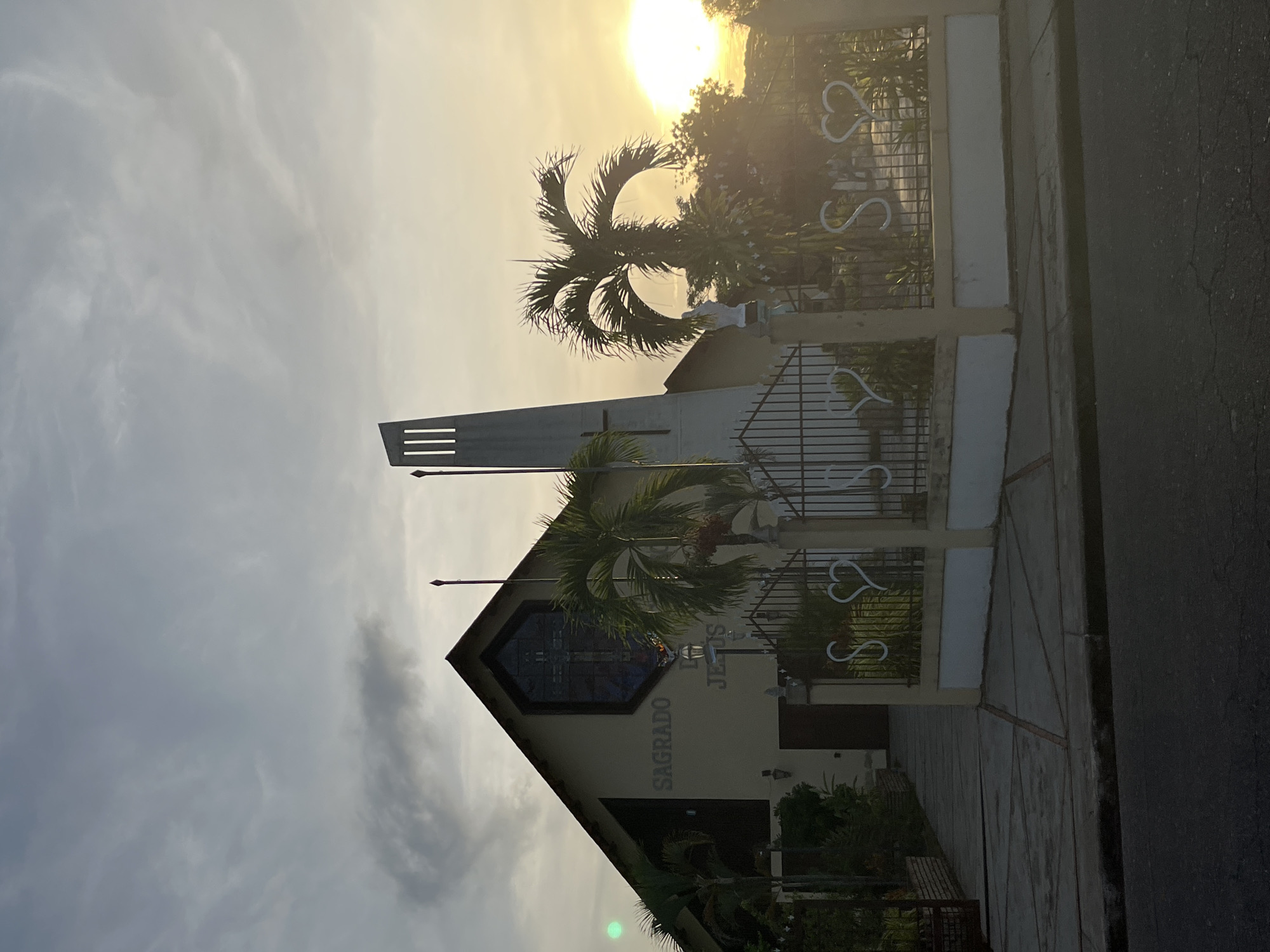 The image shows a church in Tinaquillo, Venezuela, named "Sagrado Corazón de Jesús." The building is a beige structure with a gabled roof. The facade features a stained glass window with a cross and a sign that reads "Sagrado Corazón de Jesús." A modern-style sign atop the building has horizontal slits, presumably to catch the wind. A decorative metal fence with heart shapes is in front of the building. A palm tree stands on the church grounds. The sky is cloudy with the sun shining through the clouds, creating a bright glow. Part of the street is visible in the foreground.
