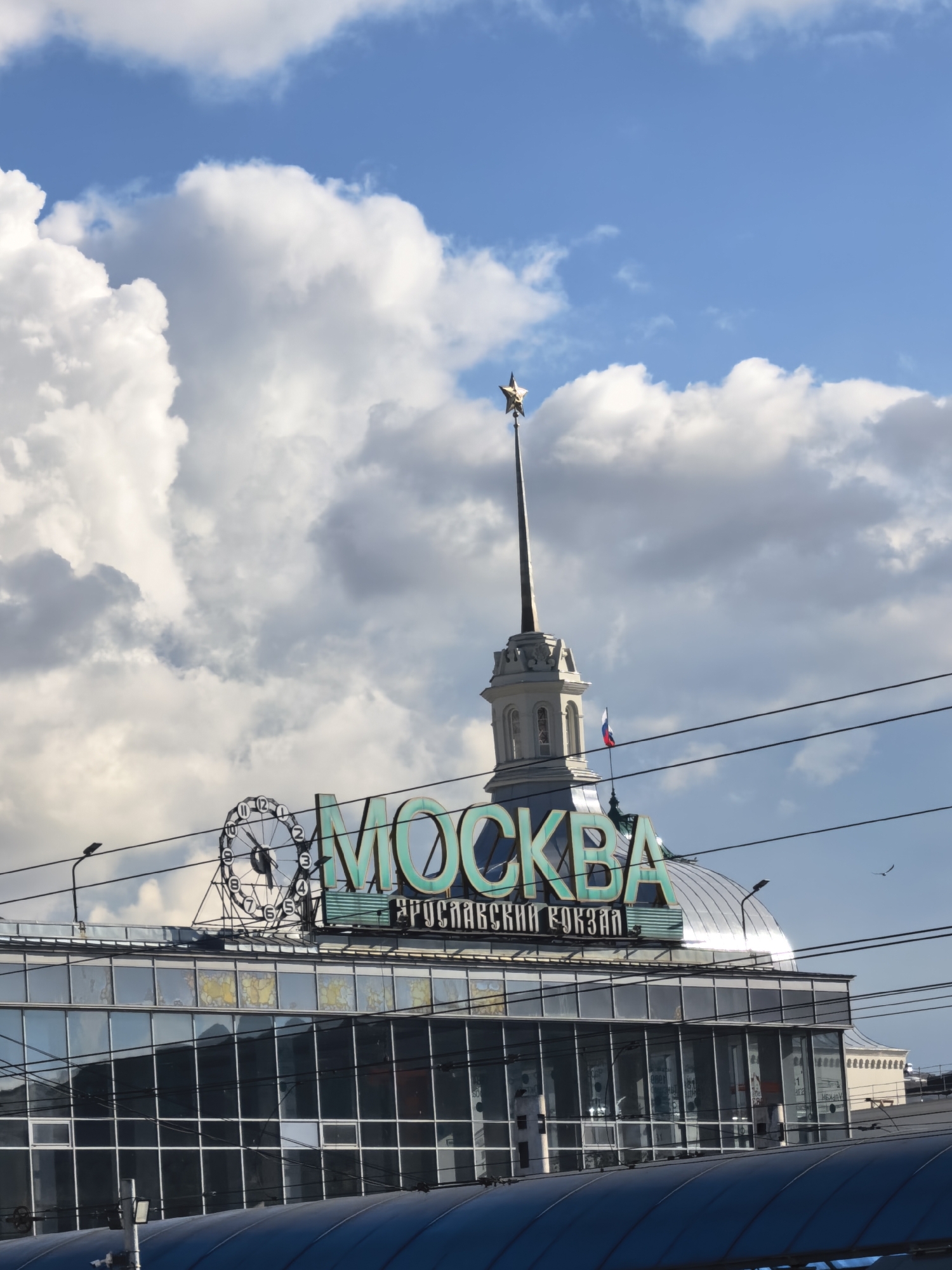 The image captures the cityscape of Moscow, Russia, specifically focusing on the Kazansky railway station. The building features a prominent spire topped with a golden star, a common architectural element in Moscow. A sign in Russian, reading "Москва" (Moscow) and "КАЗАНСКИЙ ВОКЗАЛ" (Kazansky railway station), is visible atop the building. A large clock is placed on the left part of the roof. The sky is filled with white clouds against a blue backdrop. Overhead wires cross the foreground. The building facade is primarily made of glass. A Russian flag is fixed on the building.