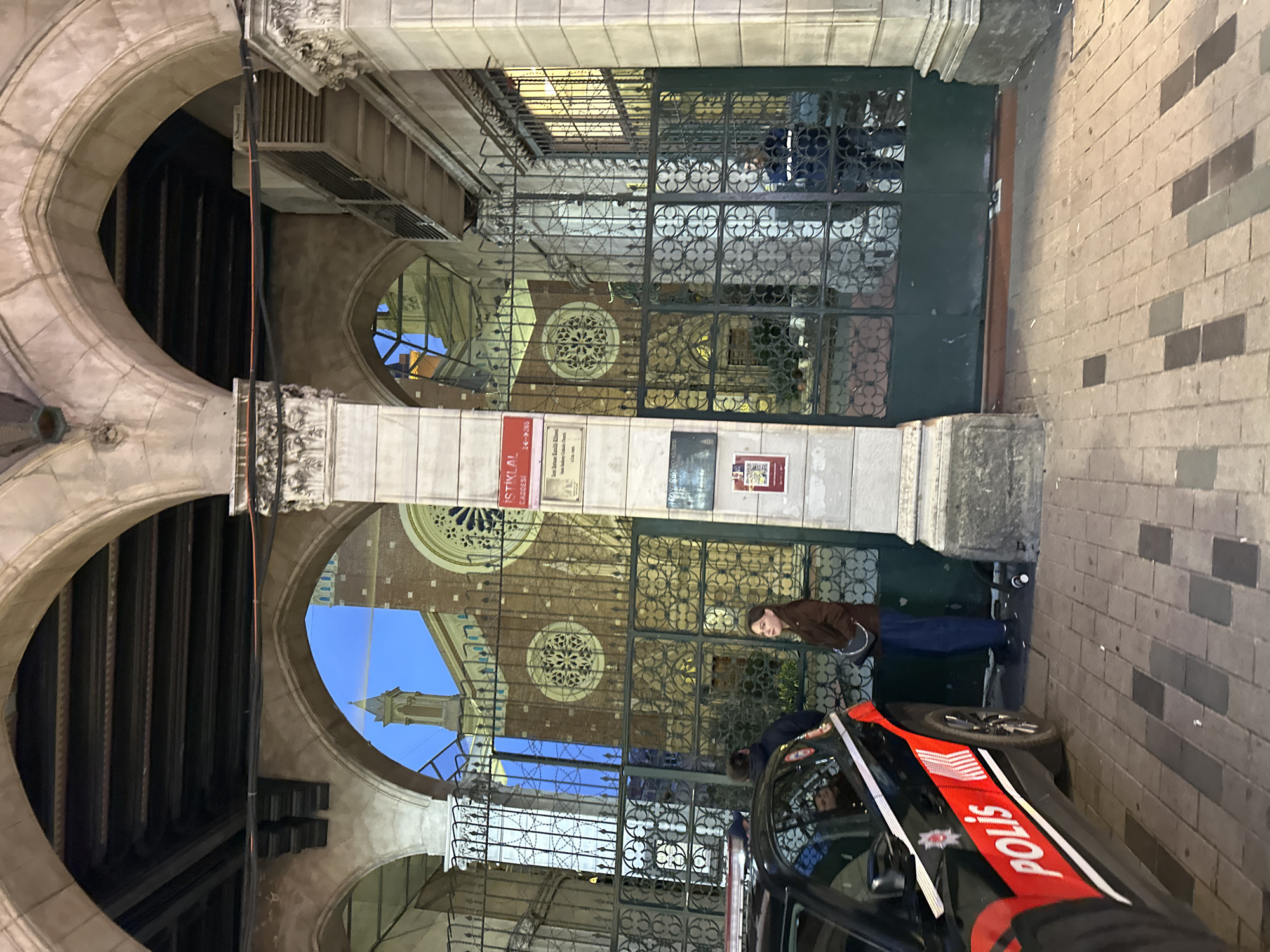 The scene depicts the arched entrance passage to the Saint Anthony of Padua Church (Sent Antuan Kilisesi) on İstiklal Avenue, Beyoğlu, Turkey, during evening hours. A black Turkish "POLIS" patrol vehicle, marked with red "POLIS" lettering and a red stripe, is parked on a brick-patterned paved surface in the left foreground. A female subject, with light brown hair, wearing a brown jacket and blue trousers, stands facing forward near the vehicle. A small white cylindrical object lies on the pavement near her. To the right, a tall, ornate dark green metal gate separates the passage from the church courtyard. This gate is situated beneath a series of stone arches supported by a central fluted column. The column displays a red street sign reading "İSTİKLAL CADDESİ 214-236" and additional plaques, one identifying the church. Through the gate, the facade of a large, reddish-brown brick church, featuring multiple rose windows and a pointed spire, is visible against a darkening blue sky. A male subject in a dark uniform is partially visible standing behind the gate on the right side. Another individual in dark clothing is partially obscured near the police vehicle on the left. An air conditioning unit is mounted above the rightmost arch.