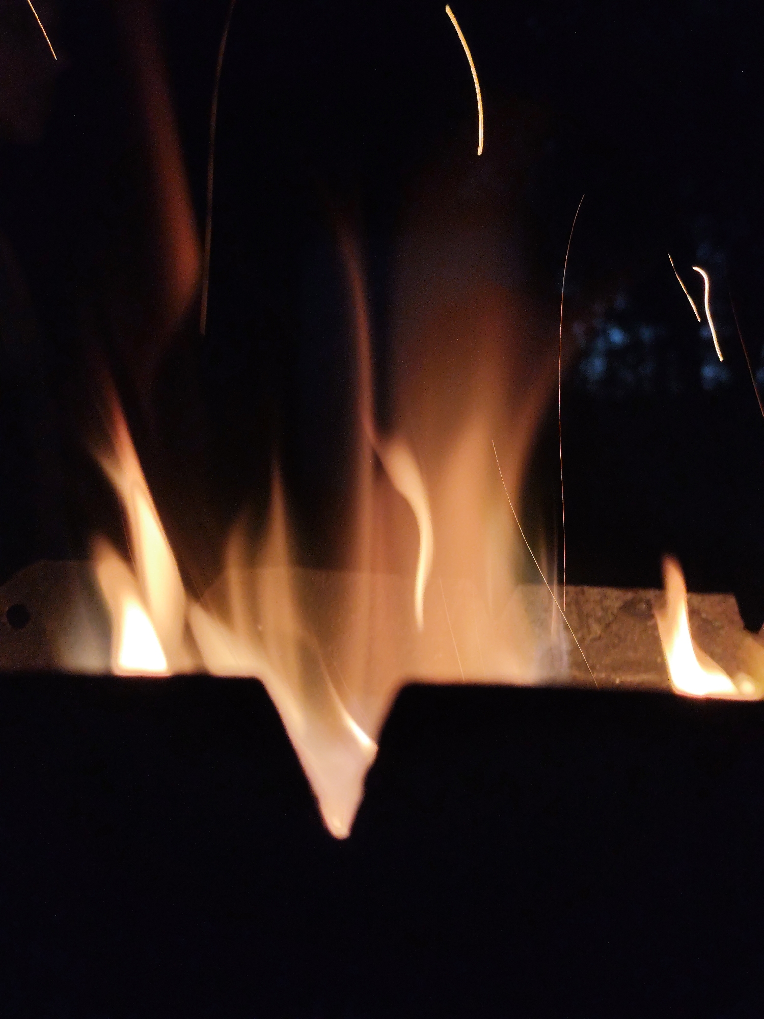 In the city of Kolpakovo, Russia, a close-up, low-angle shot captures a roaring fire at night. The flames, in shades of orange and yellow, dance against a dark background, with thin sparks ascending into the darkness. A metal grate or trough, possibly part of a brazier or fire pit, is visible at the bottom of the frame, with flames emerging from its edges. The overall impression is one of intense heat and motion within a confined space.