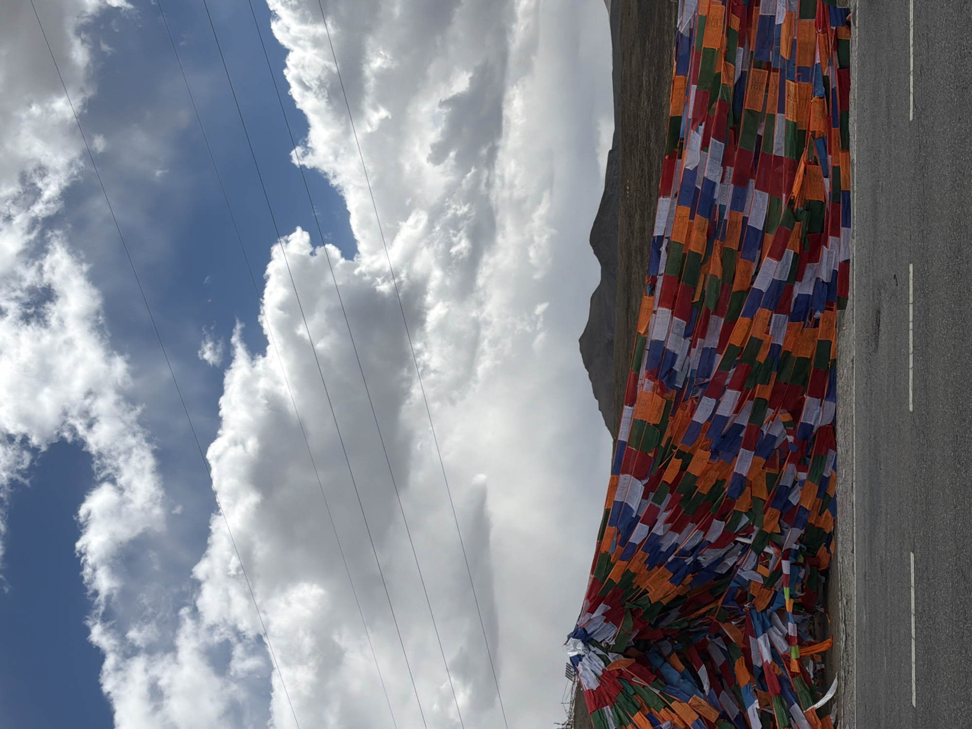 This image captures a vibrant and expansive high-altitude landscape, likely in the vicinity of Lhatsé, China. The scene is dominated by a dramatic sky, featuring patches of clear blue interspersed with large, dynamic white and grey clouds that suggest either a partly cloudy day or approaching weather. Several thin, dark power lines stretch horizontally across the upper portion of the sky.
A multitude of colorful Tibetan prayer flags, known as lungta, form a prominent visual element, strung densely across the mid-ground. These rectangular flags, in traditional colors of blue, white, red, green, and yellow/orange, are tightly packed and appear to be fluttering slightly, creating a rich tapestry of color. They extend from the left towards the right, partially obscuring a dark, rugged mountain or hillside that rises in the background. The muted tones of the mountain contrast sharply with the bright hues of the flags.
At the bottom of the frame, a paved road with distinct white dividing lines (a solid line and a dashed line) runs horizontally, suggesting a mountain pass or a highway through the terrain. The road surface appears grey and well-maintained.
The image depicts a static scene without any visible people or specific activity. The bright daylight indicates it is daytime, with the light suggesting a mid-day period. The prevalence of the prayer flags serves as a strong cultural and geographical cue, pointing to the spiritual traditions of the Himalayan region. No visible text is present in the image.