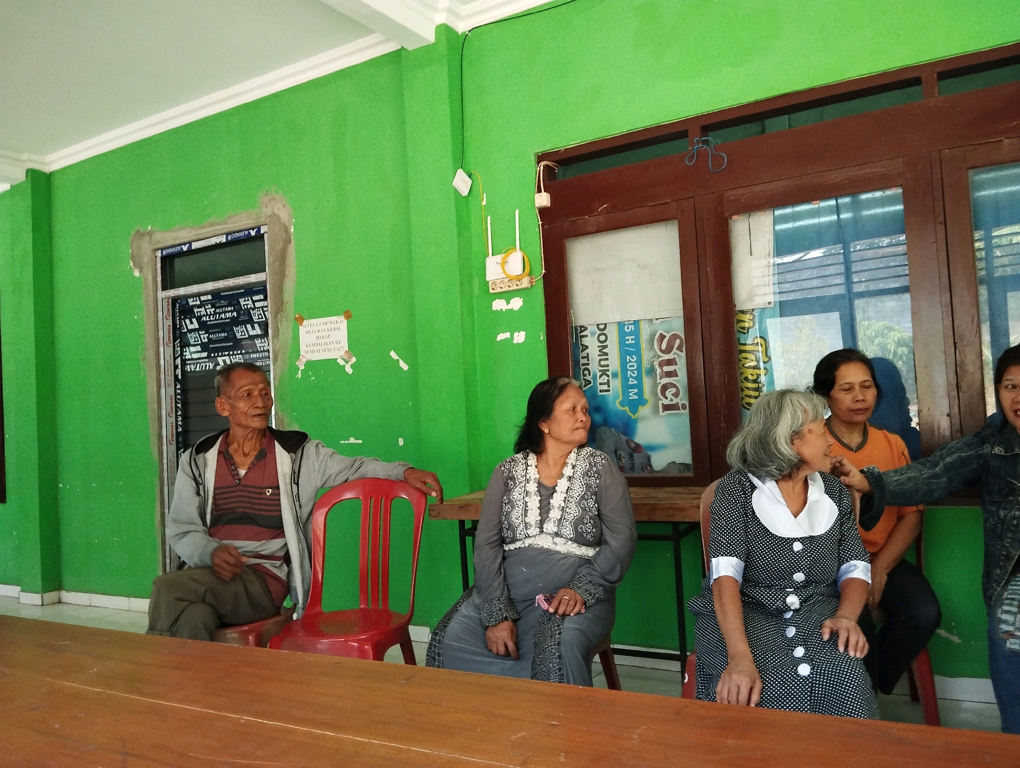 An indoor scene in the city of Salatiga, Indonesia, depicts five individuals seated at a wooden table. The room is painted a vibrant green. On the left, an elderly man with gray hair, wearing a grey jacket over a striped shirt and dark pants, is seated in a red chair. To his right, a woman with dark hair, adorned in a grey dress with white floral detailing, is also seated. Next to her, an elderly woman with grey hair wears a black and white polka-dotted dress. Behind her, another woman in an orange shirt is visible. The person furthest to the right, whose face is partially obscured by the frame, wears a denim jacket. The individuals appear to be engaged in conversation. The environment includes a large wooden table in the foreground, a double-door window with dark brown framing in the background, and a wireless router mounted on the green wall. The scene suggests a casual gathering or meeting.