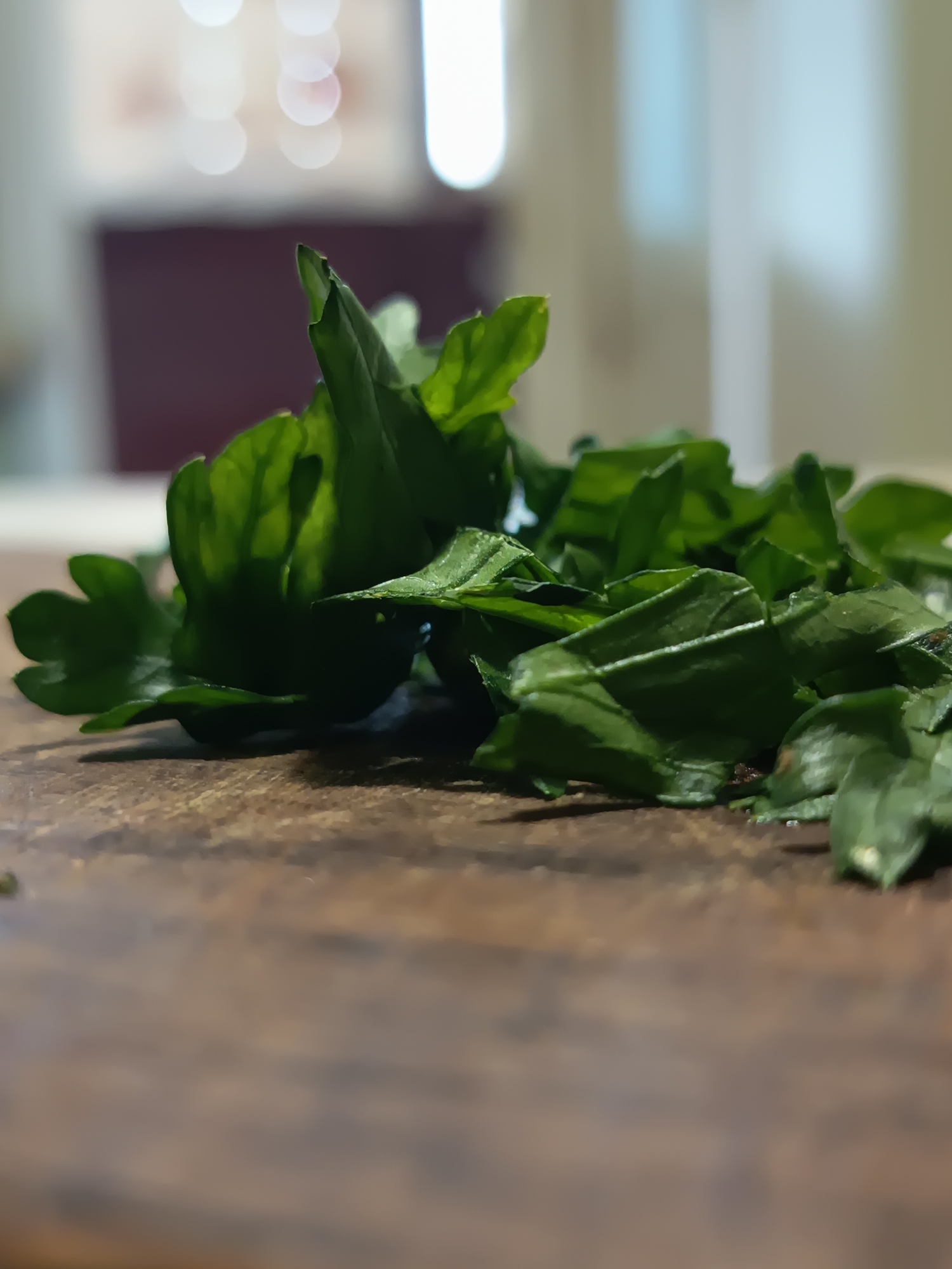 A close-up, low-angle shot captures a pile of freshly chopped parsley on a rustic wooden surface. The parsley is a vibrant green, with some leaves appearing whole and others finely diced, suggesting preparation for cooking. The shallow depth of field blurs the background, revealing soft, out-of-focus circles of light and muted colors, hinting at an indoor domestic setting. The wooden surface exhibits a rough texture and a warm, brown hue, indicative of a cutting board or tabletop. There are no people present in the image. The scene is static and peaceful. The location of Izmail, Ukraine is not discernible from the visual information provided in the image.