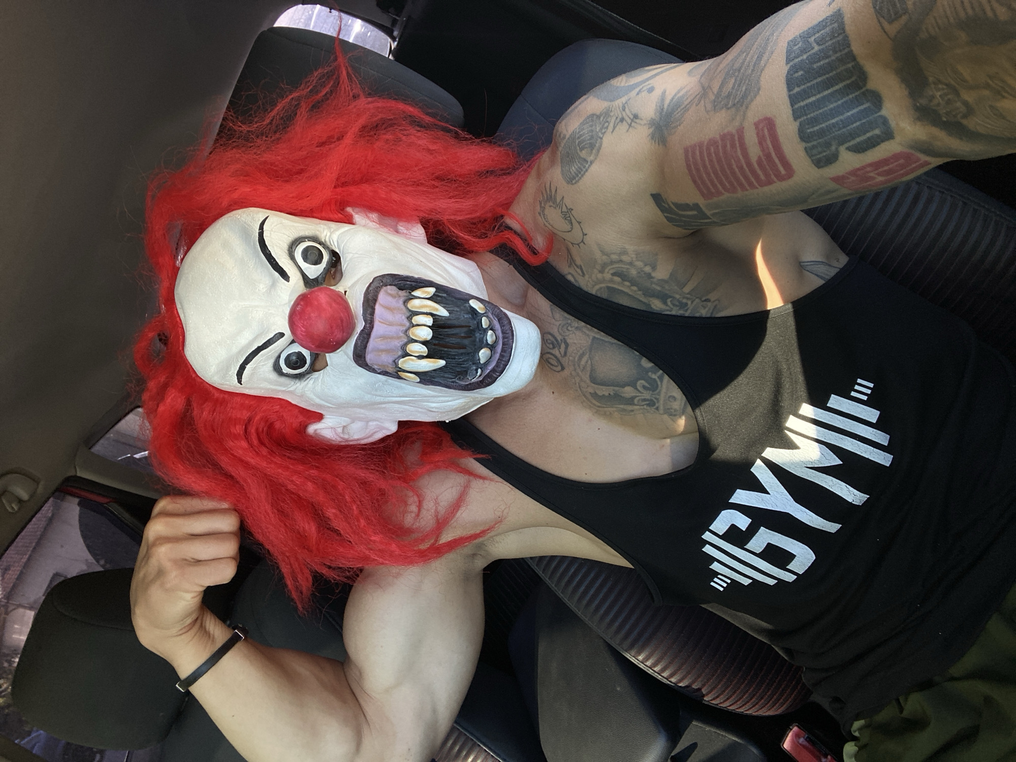 A person wearing a clown mask with red hair, a red nose, white face paint, black eyebrows, and sharp teeth, is flexing their arm inside a vehicle. The person is also wearing a black tank top with the word "GYMI" printed in white, along with imagery of weightlifting equipment. The background shows the interior of a car, including seats and windows. The image is taken from a slightly high angle, looking down at the person. There are tattoos visible on the person's arms and chest. There is no indication of the location being in the city of Pleven, Bulgaria.