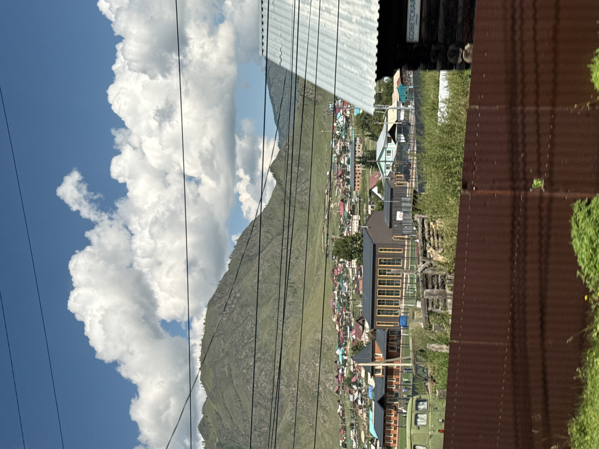 In Onguday, Russia, a village lies nestled at the foot of a lush green mountain under a bright blue sky dotted with fluffy white clouds. The village is comprised of numerous houses with colorful roofs, creating a vibrant patchwork. In the foreground, a rusty corrugated metal fence runs horizontally, partially obscuring the view of the village. Behind the fence, there are some wooden structures and a green vehicle. Further back, a dark wooden building with a corrugated metal roof is visible on the right side of the frame. The overall scene depicts a rural residential area in a mountainous region.
