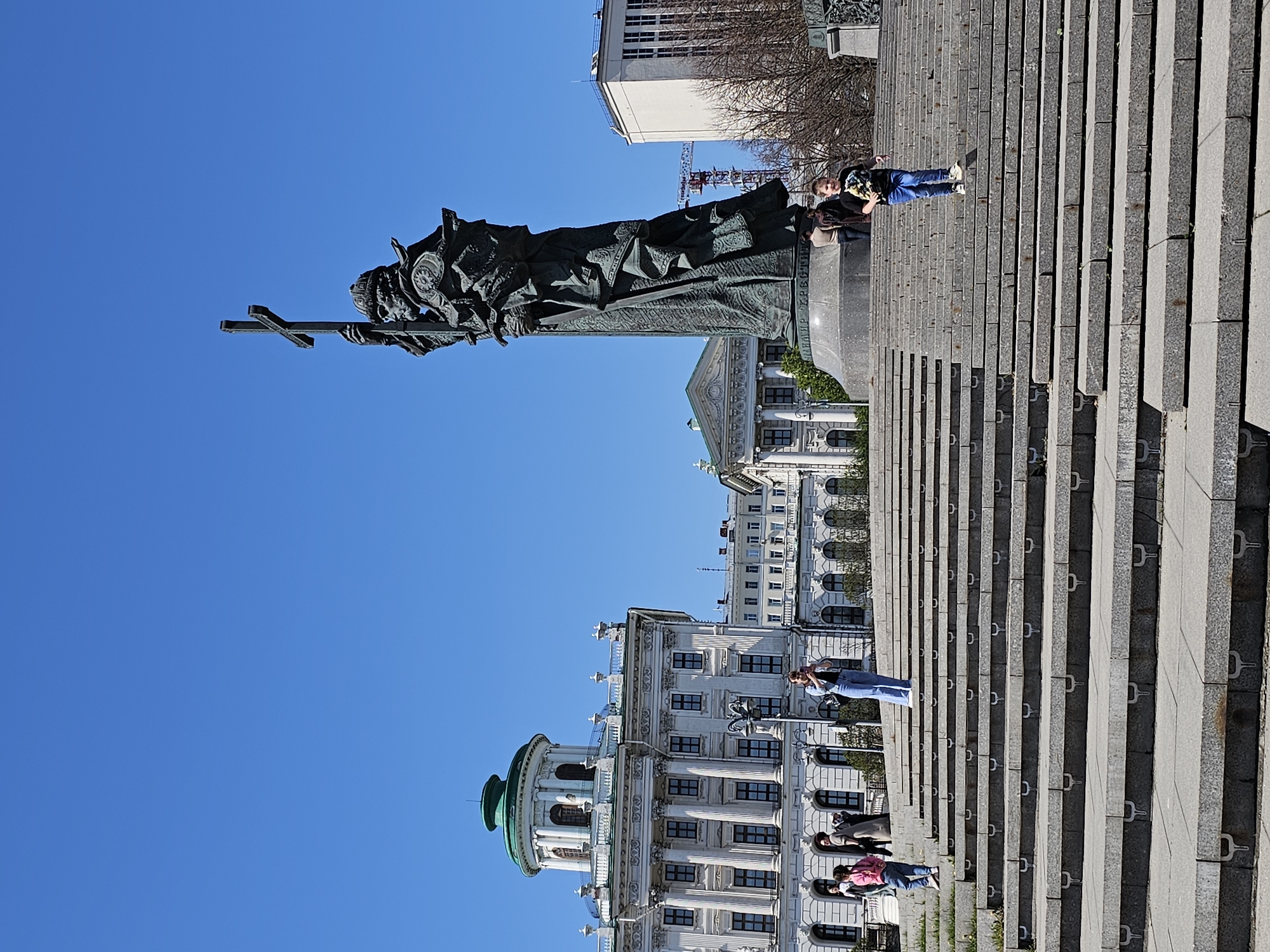 Here's a detailed description of the image:
**Location:** The image captures a scene at the Monument to Saint Vladimir on Borovitskaya Square, Moscow, Russia. The buildings in the background suggest a central location.
**Subjects and Objects:**
* A large bronze statue of Saint Vladimir holding a cross is the central focus. The statue is detailed with intricate drapery.
* Several people are present on the wide staircase leading up to the monument. They are positioned at different levels of the stairs.
* The staircase is made of gray stone and has metal fixtures on each step.
* Buildings with architectural detail, including rounded domes and classical features, are visible in the background. Some have windows and architectural details.
* Clear blue sky is overhead.
**People and Actions:**
* At least three people are sitting on the stairs. One person is near the base of the monument. Another is in the background.
**Environment:**
* The weather appears clear and sunny.
* The scene is urban, with large buildings and a major monument.
* Some trees are visible in the background.
