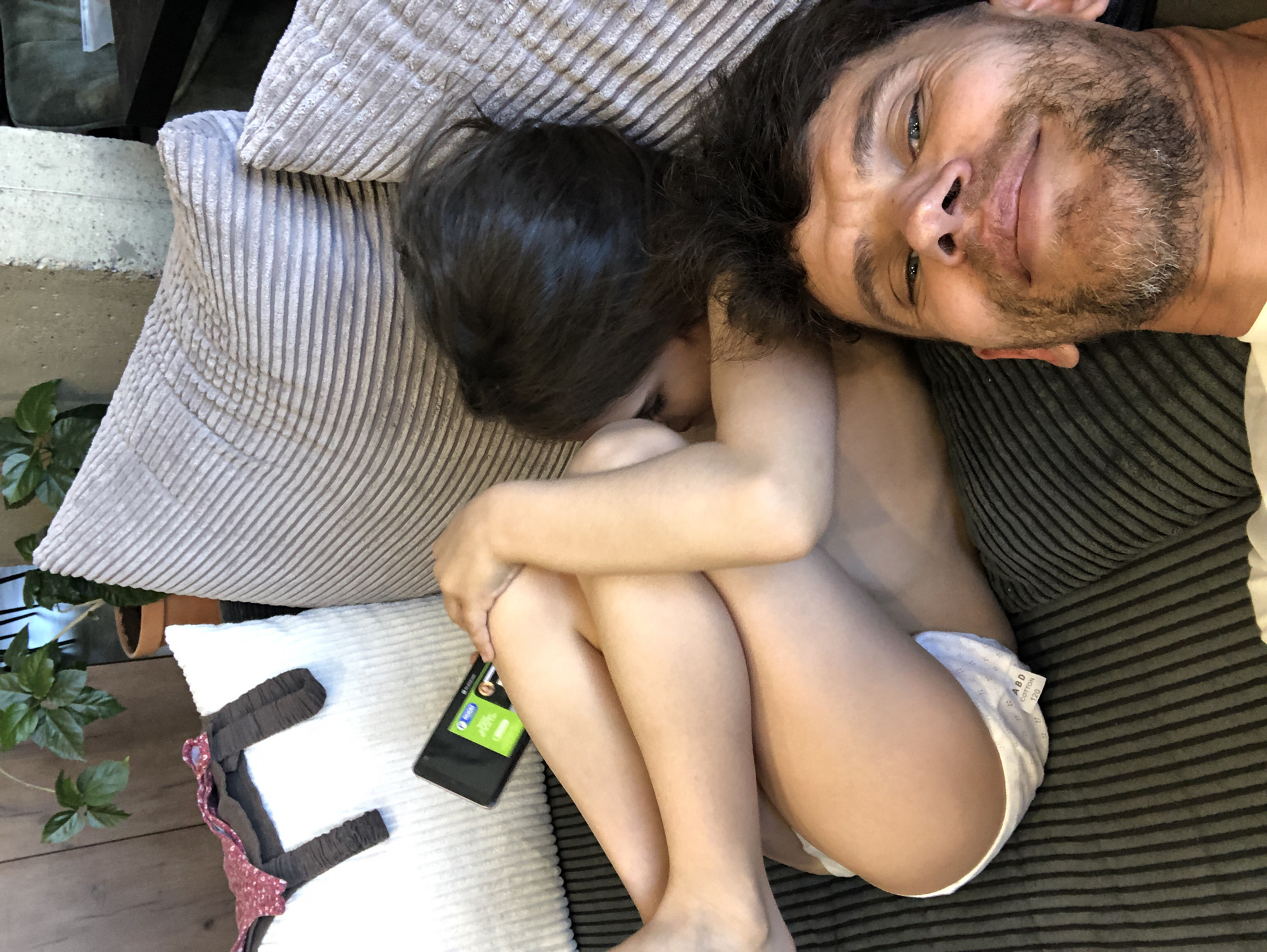 An adult male is positioned in the foreground, taking a selfie in an indoor, domestic setting. He has short dark hair with visible grey, a short beard with grey, and blue eyes. Behind him, a child is curled up on a sofa, facing away from the camera, with dark hair visible. The child is holding a dark smartphone with a brightly lit green screen and is wearing white briefs with a visible label reading "ABD COTTON 120". The sofa features ribbed fabric upholstery in light grey on the backrests and darker grey on the seating area. A large white ribbed pillow is on the left, along with other light grey ribbed pillows. On the white pillow, a reddish-pink patterned fabric item and a dark brown ribbed fabric item are present. The background reveals wood paneling, a potted green plant in a terracotta pot, a concrete structural element, a dark table, and a partially visible green upholstered chair.