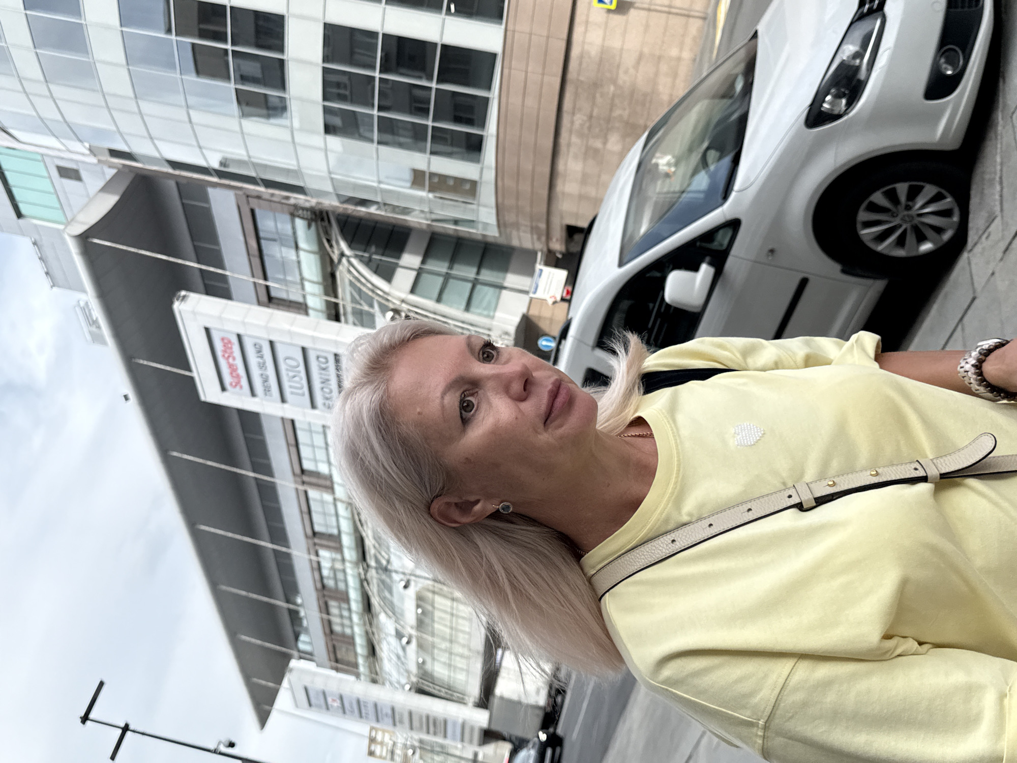 A light-skinned woman with shoulder-length blonde hair, wearing a pale yellow t-shirt and a crossbody bag, walks along a city sidewalk. She is looking off to the right of the frame. Behind her, a white van is parked on the street. In the background, a modern building with a glass facade and a prominent canopy with multiple signs, including "SuperStep," "TREND ISLAND," "LUSIO," and "EKONIKA," rises against an overcast sky. The location appears to be Moscow, Russia, given the signage and architectural style.