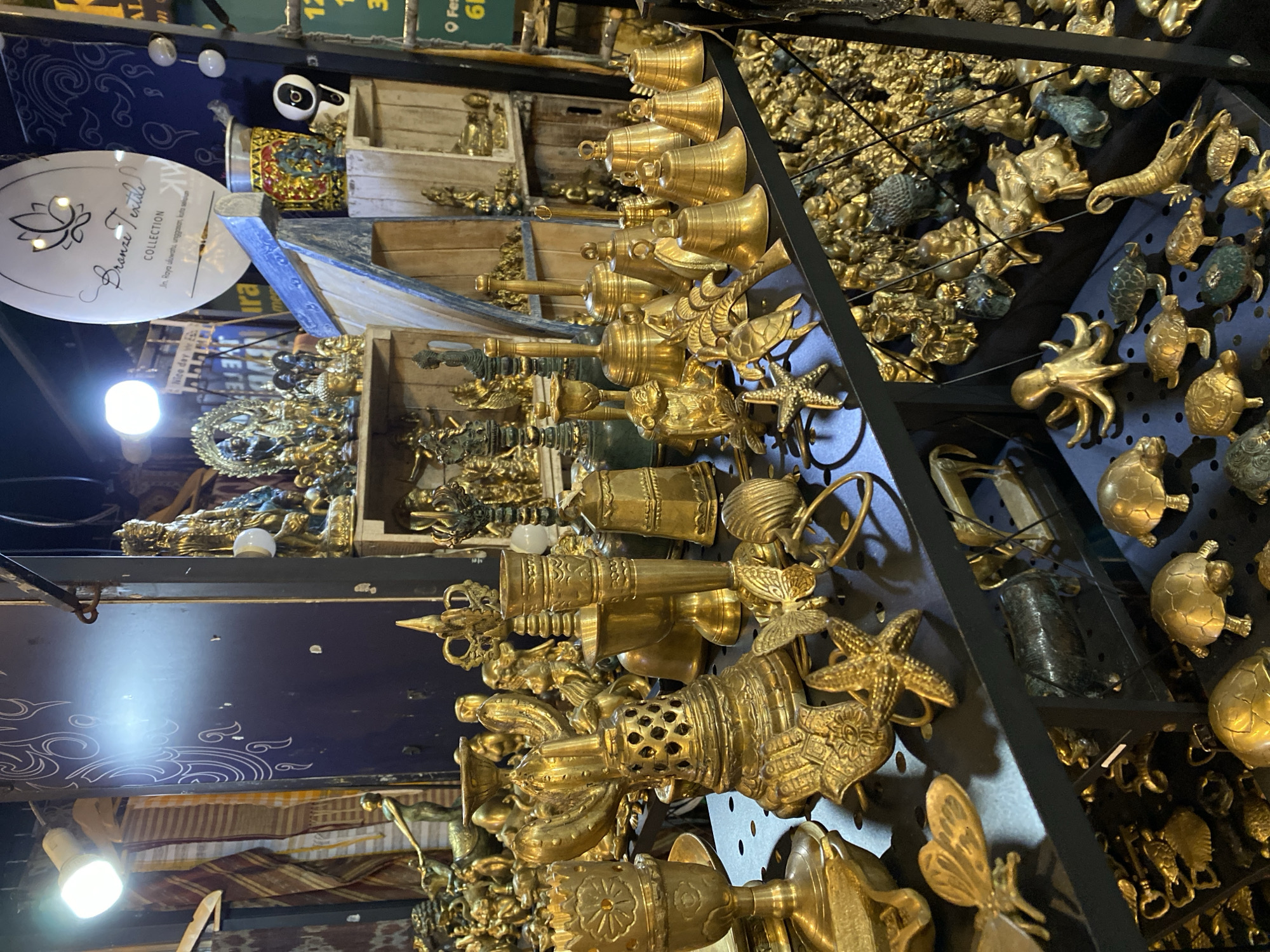 The image depicts a stall in Kuta Selatan, Indonesia, selling a variety of brass figurines and decorative items. The stall is filled with numerous golden brass objects, including small statues of deities, animals such as turtles, starfish, elephants, and octopuses, as well as bells and ornate candle holders. Some of the items are arranged on shelves, while others are displayed on tables. In the background, there are textiles and a sign that reads "Bronze Textile Collection". A surveillance camera is visible on the right side of the image. The lighting suggests an indoor market or shop. There are no people visible in the frame.