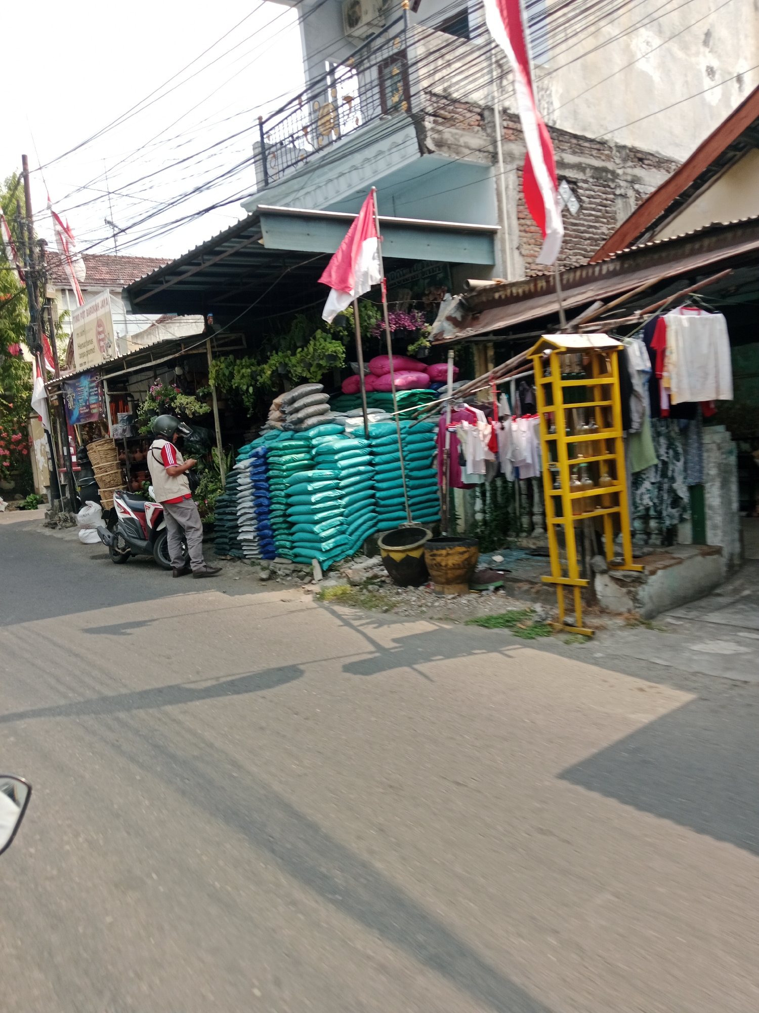 The image depicts a street scene in Kediri City, Indonesia, with a man standing next to a motorcycle. He is wearing a red and white vest, grey pants, and a helmet. The motorcycle is red and white. In the background, there is a building with a dark awning and various goods displayed, including stacks of green and blue bags, pink bags, baskets, and clothes hanging on a rack. A yellow rack with bottles is also visible. Indonesian flags are displayed on poles. The street in the foreground is paved, and shadows from overhead wires and structures are cast across it.