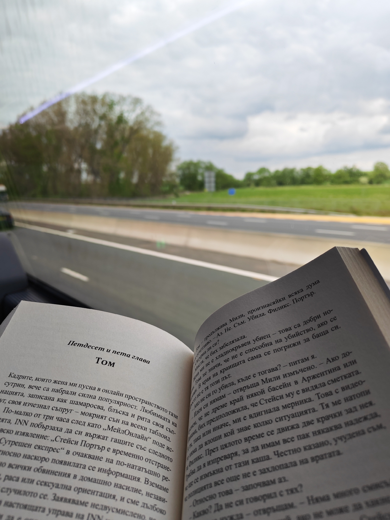 A person is reading a book while apparently traveling on a highway, as evidenced by the passing scenery through the car window. The book is open to two pages with text in Bulgarian, titled "Петдесет и пета глава Том" ("Fifty-fifth Chapter Tom").
Outside the car window, the landscape consists of trees and green fields under an overcast sky. The highway appears to have multiple lanes. Based on the style of the road infrastructure and signage, the location is likely the M1 motorway near Milton Keynes in the United Kingdom.