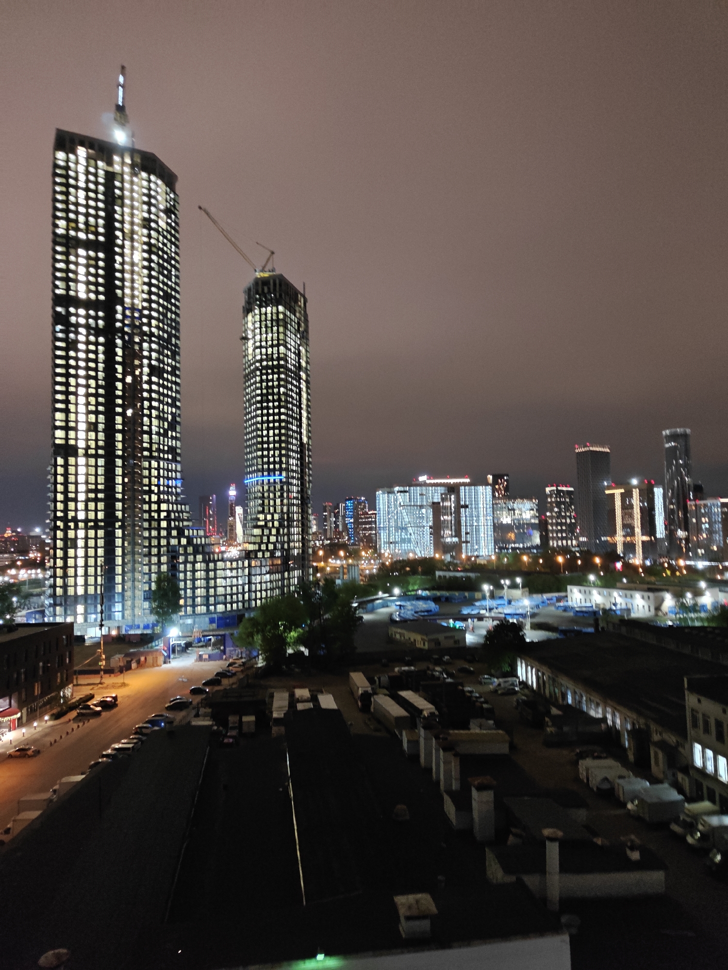 The image shows an urban cityscape at night, with the Moscow International Business Center (Moscow City) prominent in the background. Two large skyscrapers dominate the foreground, both lit with numerous windows, one seemingly still under construction with a visible crane. Below the skyscrapers lies an industrial area, with long buildings and what appear to be parked trucks. A street lined with parked cars is visible to the left. The sky is overcast and dusky.