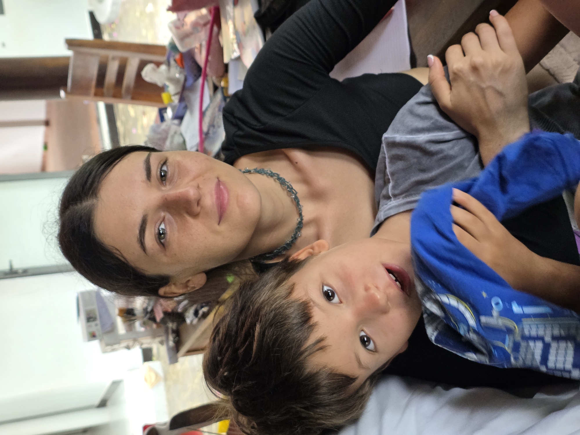 An intimate, candid portrait captures a woman and a young child lying down together, both looking directly at the camera. The image is oriented vertically, showing the woman on the left and the child on the right, suggesting they are resting or posing on a bed or similar surface.
The woman has dark hair pulled back, fair skin with some freckles, and light brown eyes that engage with the viewer. She displays a soft smile and wears a black long-sleeved top along with a distinctive grey-blue beaded necklace. Her hand, with light-colored nail polish, is visible, gently resting near the child.
Beside her, the child, with brown hair and wide, dark eyes, also looks towards the camera. The child's mouth is slightly open, revealing teeth, hinting at a playful or curious expression. The child is dressed in a grey t-shirt layered with a blue t-shirt featuring a subtle graphic design.
The background is softly blurred, indicating an indoor setting with ambient light, likely during the daytime. Out-of-focus elements suggest a casual, domestic environment, possibly a room with some furniture, such as a wooden chair or table, and various personal items. The overall mood of the photograph is warm and affectionate, capturing a tender moment between the two. The scene is set in Mũi Né, Vietnam. No discernible text is visible in the image.