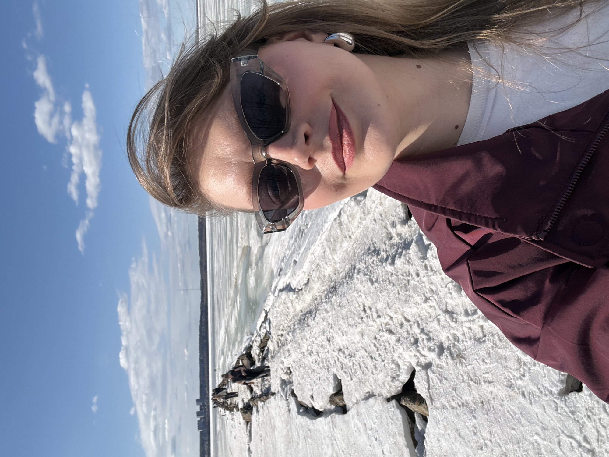 A woman is standing on a snow-covered shoreline in Izhevsk, Russia. She wears sunglasses, a white t-shirt, and a maroon jacket. Behind her is a body of water, likely a lake or reservoir, partially covered with ice. The sky is blue with scattered clouds. The shoreline has a rough texture due to the snow and ice. A cityscape is visible in the background across the water.