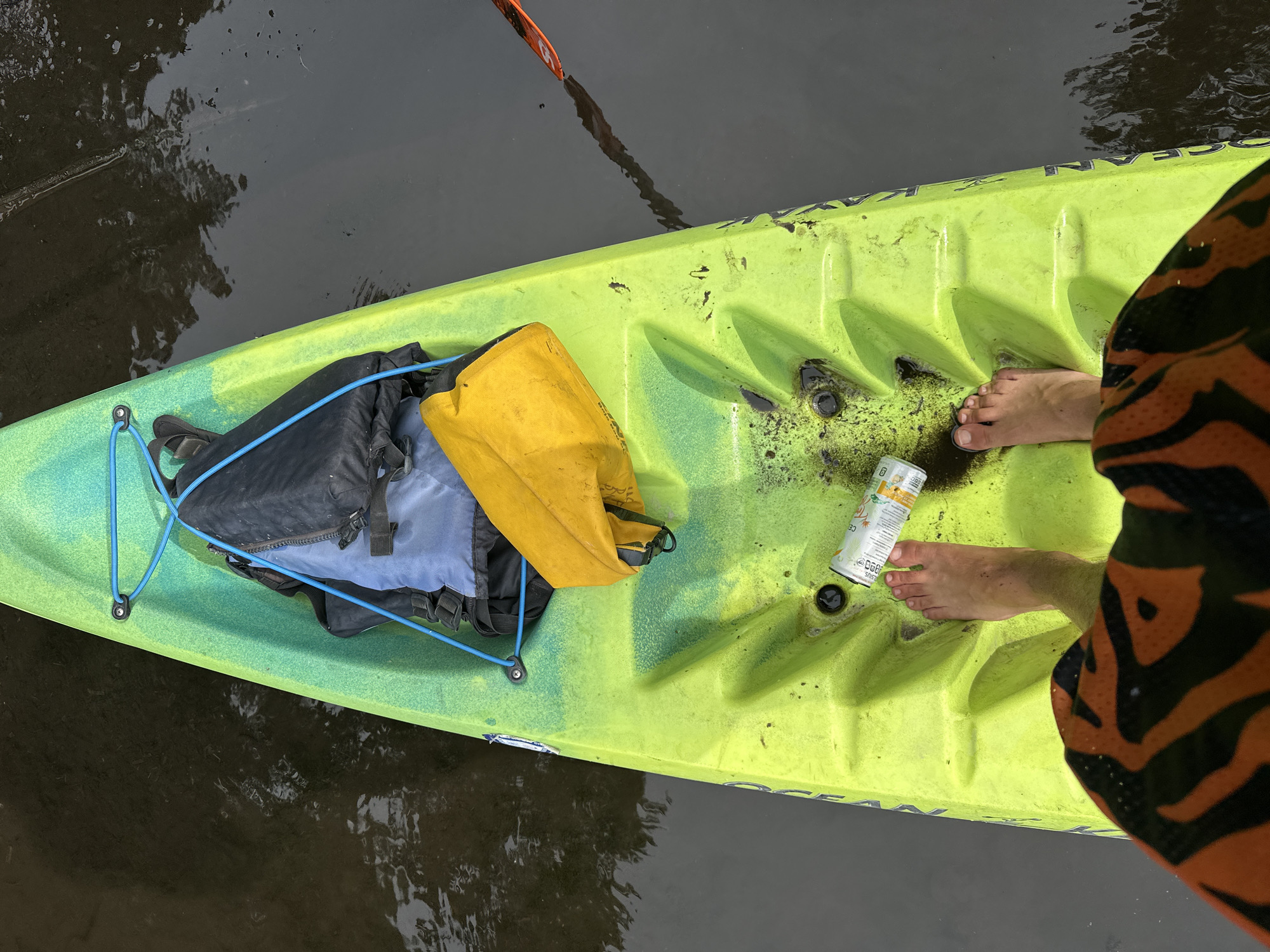 An overhead shot captures a pair of bare feet inside a bright lime green and yellow kayak, floating on dark, calm water. The kayak appears to be a sit-on-top model, with the brand name "OCEAN KAYAK" partially visible along the side. The interior of the kayak is marked with some dark dirt and debris, particularly around the foot area.
Resting in the front of the kayak is a black and blue backpack, secured by a bright blue bungee cord system. Next to the backpack is a yellow dry bag. Near the person's feet, a white beverage can with colorful graphics is visible, displaying the text "CELSIUS", "ESSENTIAL ENERGY", and "ZERO SUGAR". The can also indicates "GREEN TEA &..." and other small, illegible print.
The person's feet are positioned towards the rear of the kayak, and the bottom of their orange and black tiger-print swim shorts is visible on the right side of the frame. A portion of an orange kayak paddle blade is seen in the water, just beyond the front of the kayak. The dark water reflects distorted images of trees and sky, suggesting an outdoor setting like a river or calm lake. The scene depicts a moment of rest during a kayaking activity in Tampa, United States.