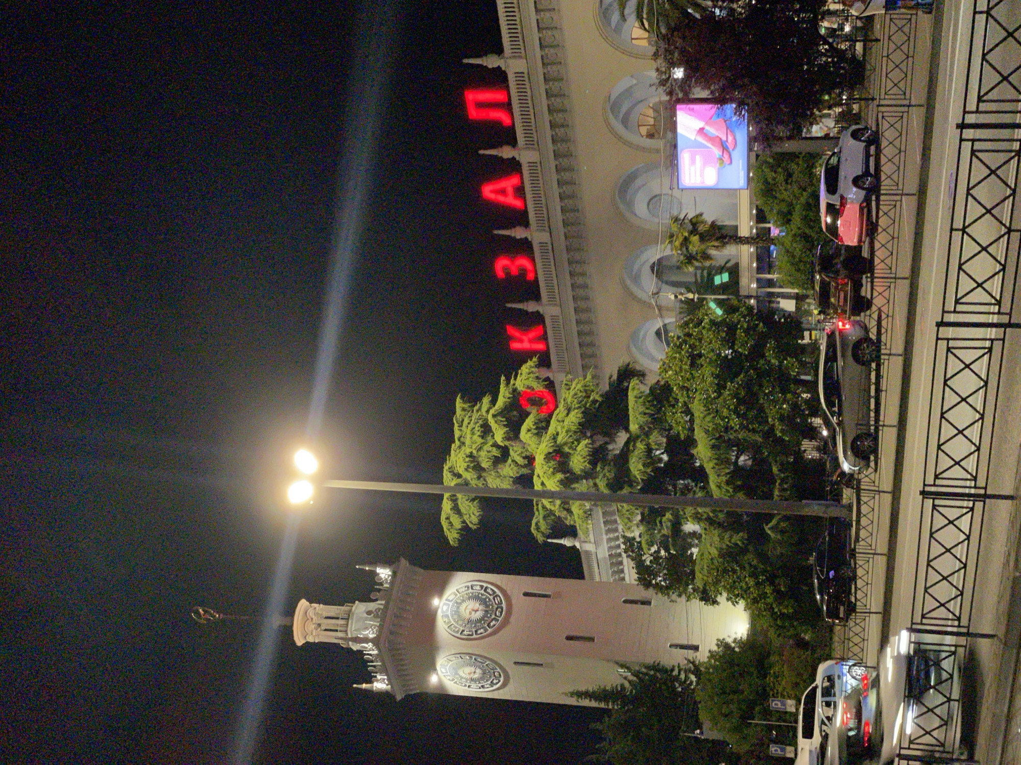 This nighttime photograph captures the exterior of a building in Sochi, Russia, identified by the illuminated red lettering on its facade spelling out "КАЗАЛ" which likely refers to a train station or similar public building. To the left of the building, a tall, multi-tiered clock tower with illuminated clock faces stands prominently. The tower is topped with a decorative spire. In front of the building, a row of parked cars is visible, and a street with traffic is in the foreground, separated by a decorative black metal fence. Large, lush trees, including evergreens and palms, are interspersed throughout the scene, providing a contrast to the urban architecture. A large digital billboard displaying an advertisement featuring a close-up of feet wearing sandals is mounted on the building's exterior. The sky is dark, indicating it is night, and a bright streetlight illuminates the immediate area.