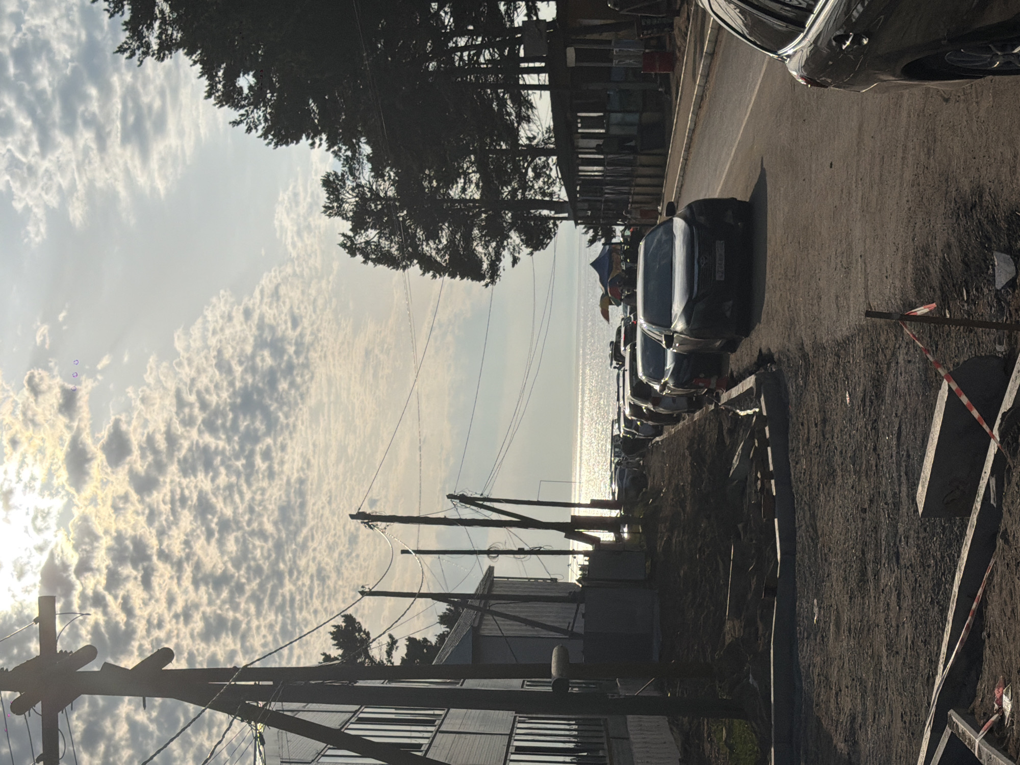 The image shows a street scene in Goryachinsk, Russia, with a body of water in the background. The foreground is dominated by a street under construction, with piles of dirt and construction materials. A line of cars is parked along the side of the street, and a few people are visible walking or standing near a tented area. Tall pine trees line the right side of the street, and a building with several windows is on the left. Utility poles with wires are scattered throughout the scene. The sky is partly cloudy, with sunlight filtering through the clouds.