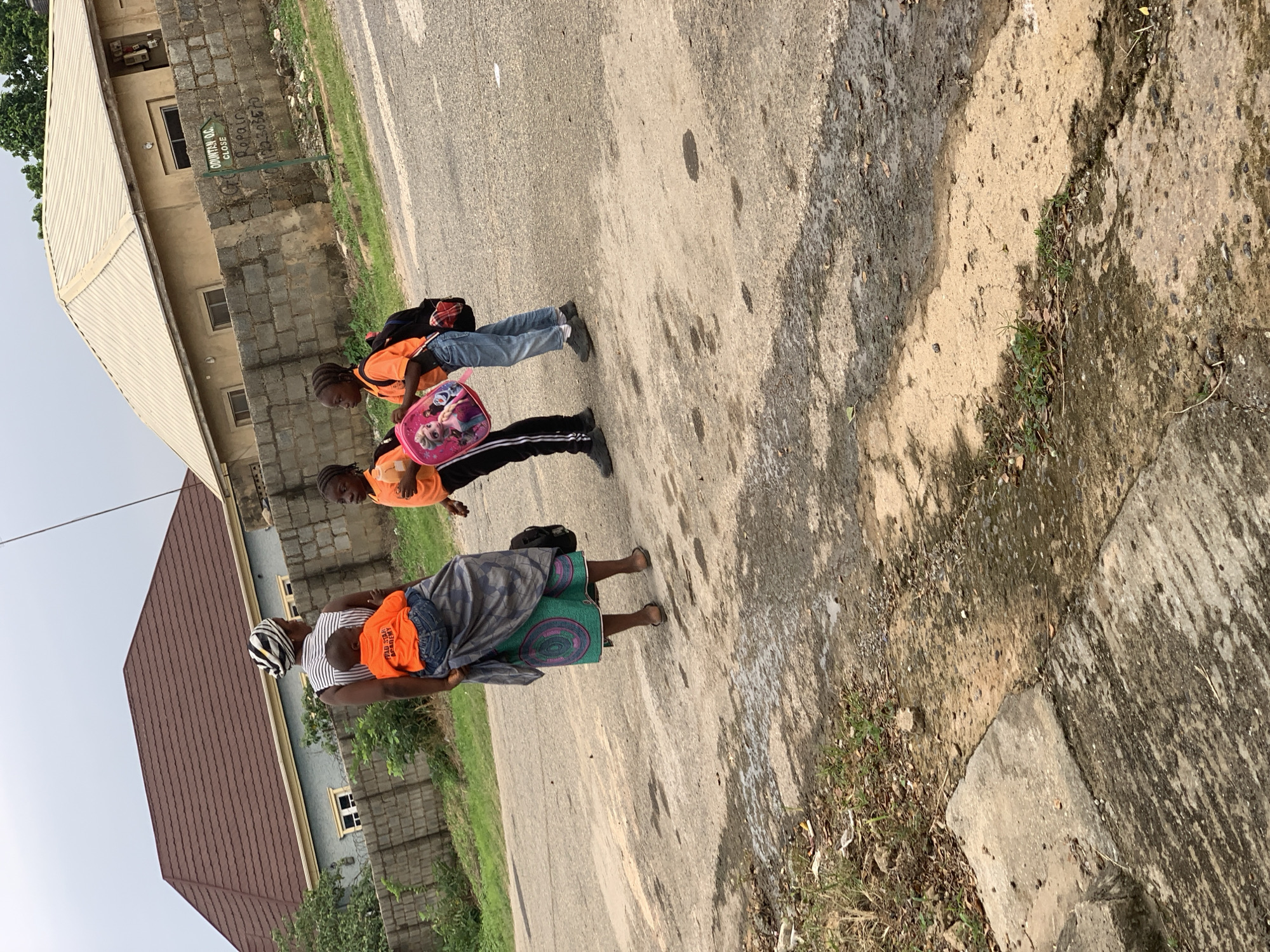 A woman with a striped headscarf is carrying a baby on her back while walking on a paved road. She is wearing a striped top and a colorful wrap skirt. Two young children, a boy and a girl, are walking beside her, both wearing orange shirts and blue jeans. The girl is carrying a pink backpack with images of cartoon characters, and the boy is carrying a black backpack. They appear to be on their way to or from school. In the background, there are buildings with brown and beige roofs, a brick wall, and lush green trees. The sky is overcast. A directional sign with text "ODUNTAN O.C CLOSE" is visible on the brick wall. The overall setting suggests a residential area within Abuja, Nigeria.