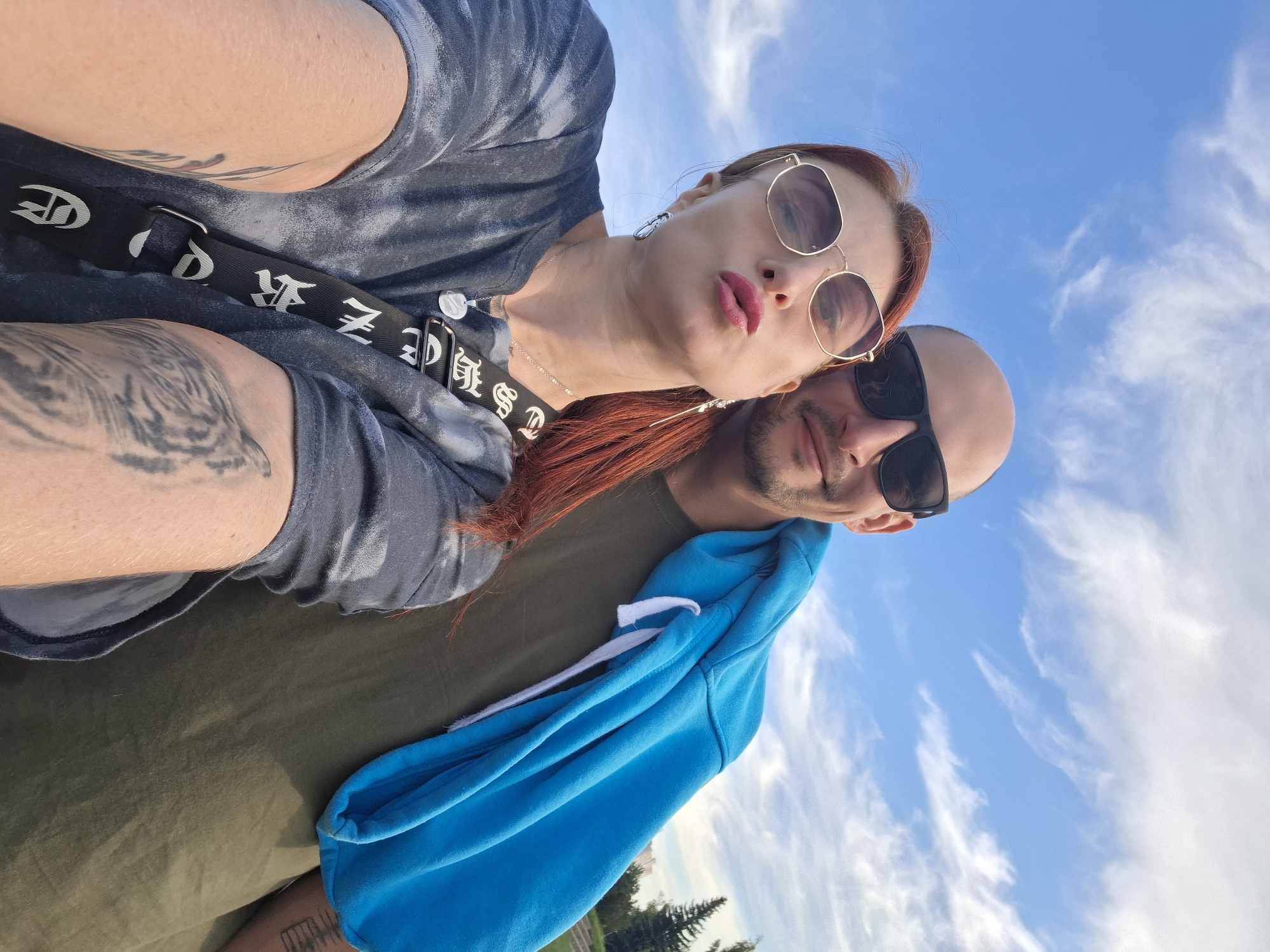 A medium shot, selfie-style photo captures a couple standing outdoors against a bright blue sky with wispy clouds. The woman, on the left, has reddish-orange hair and is wearing large, hexagonal sunglasses and a grey, tie-dye t-shirt. She is puckering her lips and wearing a necklace with a circular pendant and a black crossbody bag with white lettering. She has tattoos on her left arm. The man, on the right, is bald and wearing sunglasses and an olive-green t-shirt. He has a blue hooded sweatshirt draped over his shoulder. The background includes trees and a building. The location appears to be a park or open area in the city of Myadzyel, Belarus.
