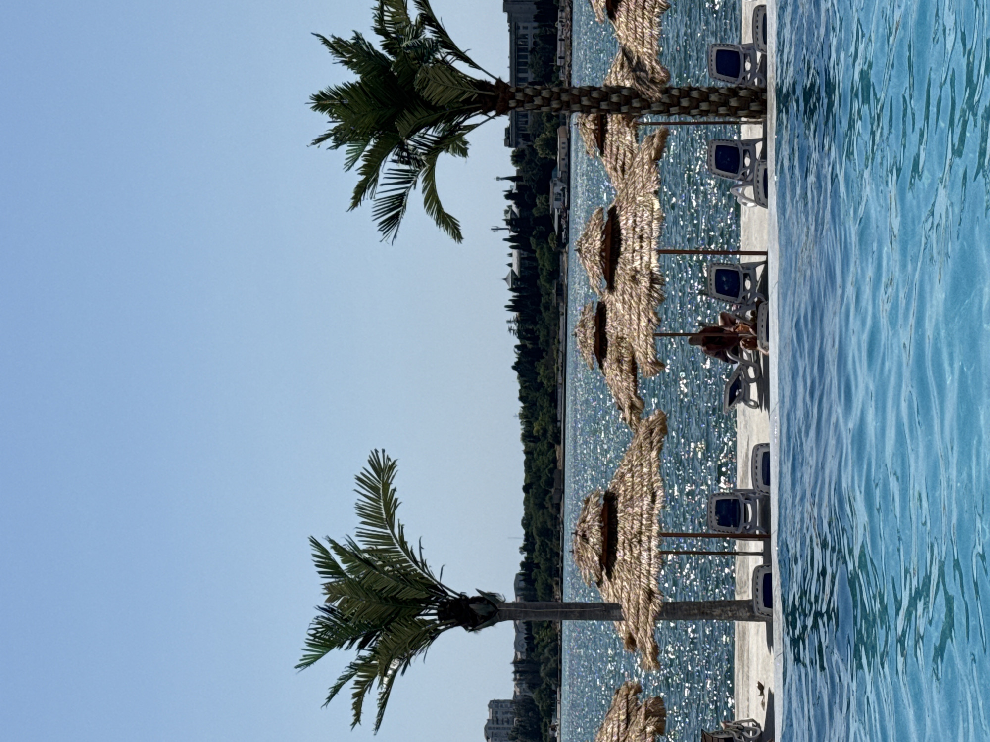 A serene, sun-drenched beach scene unfolds with a clear blue sky above. In the foreground, a shimmering swimming pool occupies the lower portion of the frame, its tranquil blue water inviting relaxation. Beyond the pool, a paved walkway lines the shore, adorned with several thatched-roof umbrellas providing shade. Beneath the umbrellas, rows of empty lounge chairs are neatly arranged, awaiting visitors. Two tall palm trees stand sentinel, their fronds reaching towards the sky, adding a tropical ambiance to the setting. The sparkling sea stretches out in the mid-ground, its surface glinting under the sunlight. In the distance, a lush green treeline forms a backdrop, beyond which a few buildings are visible, hinting at a coastal town. A solitary figure is seated on one of the lounge chairs under an umbrella, enjoying the peaceful atmosphere. The overall impression is one of a tranquil resort, a perfect escape for leisure and enjoyment.