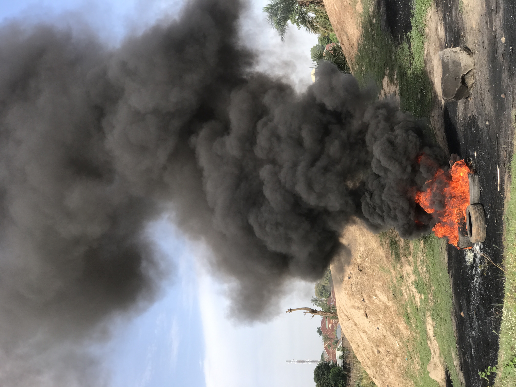 The image captures an outdoor scene dominated by a large, intense fire producing voluminous, thick black smoke under a bright, clear blue sky in Abuja, Nigeria.
In the foreground, two tires are engulfed in vivid orange flames, serving as the source of the heavy, dark grey to black smoke that rises and spreads upwards, partially obscuring the sky. The ground immediately surrounding the burning tires is charred and blackened, with some white ash-like residue visible. Adjacent to the burnt area, there are patches of green grass interspersed with small white flowers, providing a stark contrast to the destruction. A large, rough dark grey rock or concrete block sits near the grassy area.
Further into the midground and background, the landscape consists of a sloping earthen mound with sparse green vegetation. Beyond this, residential buildings with varying rooftops, numerous trees including tall palm trees, and what appears to be a communication tower are visible in the distance, suggesting a populated area.
The primary event depicted is the burning of tires, creating a significant plume of air pollution. The scene appears to be taking place during daylight hours. No people are visible in the image, nor is there any discernible text. The overall impression is one of an uncontrolled outdoor burn.
