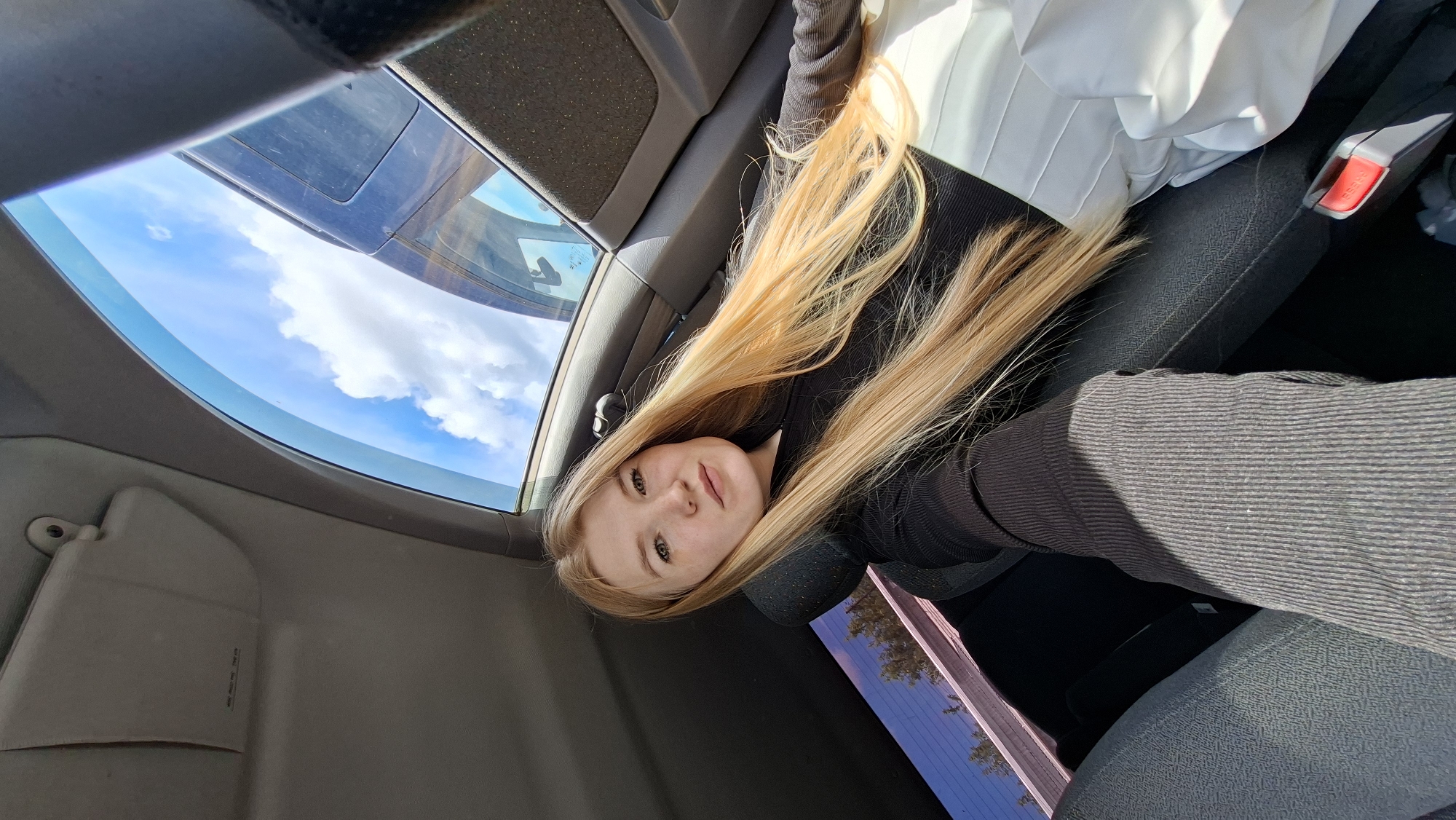 A young woman with long, blonde hair is sitting in the passenger seat of a car. She is wearing a black long-sleeved shirt and white pants. The car's sunroof is open, and a bright blue sky with white clouds is visible above. The woman is looking at the camera and smiling. The car appears to be parked, with trees and buildings visible through the rear window. The image was taken in Chaikovsky, Russia, on a sunny day.