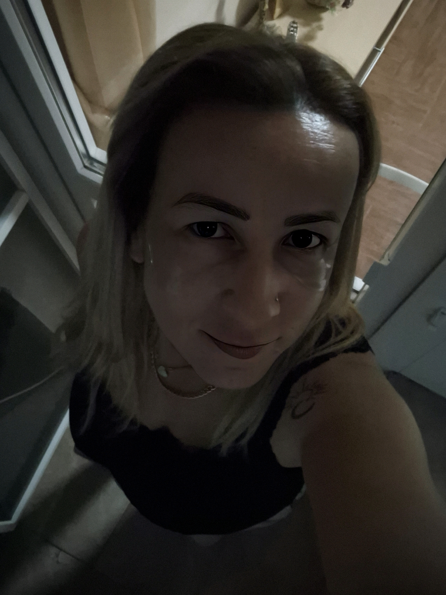 A woman is taking a selfie from a high angle, looking up at the camera. She has blonde hair, fair skin, and dark eyebrows. She is wearing a black top and a gold necklace. A tattoo is visible on her right shoulder. The background appears to be indoors, with wooden flooring and a window or doorframe. The lighting is dim, casting shadows on the scene.