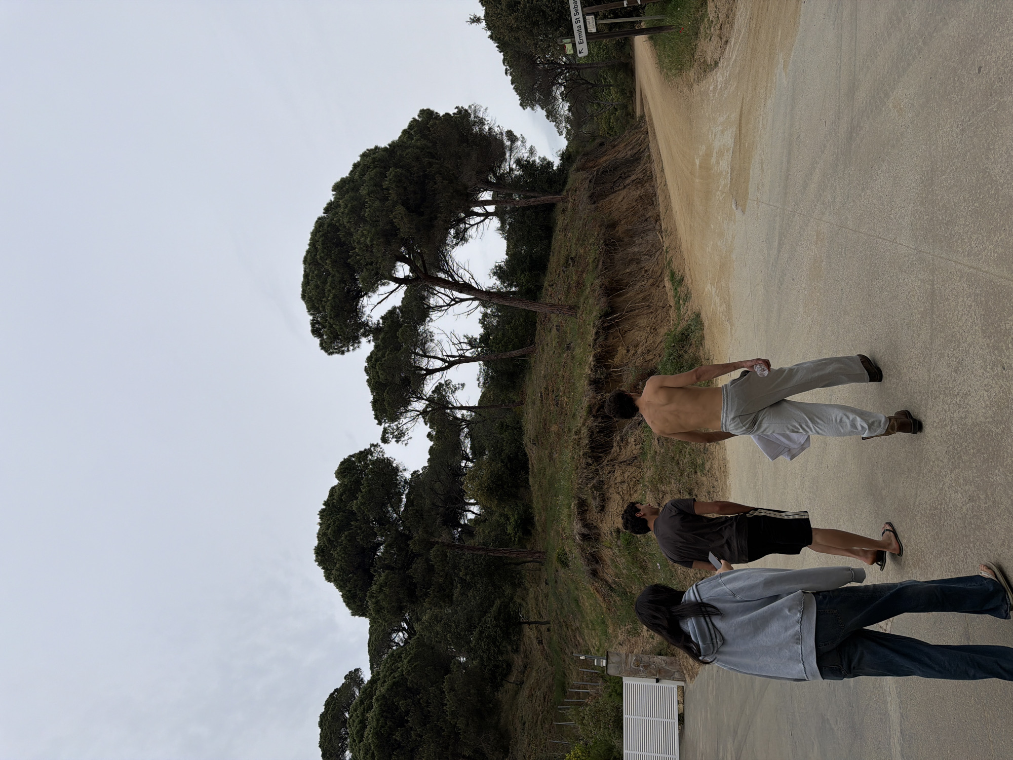 The image depicts three people on a paved path next to a dirt road, near the city of Sant Andreu de Llavaneres, Spain. A person with bare torso walks ahead, carrying a water bottle in one hand. The other two people are behind them and are dressed in casual wear. The path borders a grassy embankment with tall, dense trees. A signpost indicating "Ermita St. Sebastià" is located further up the dirt road. The sky is overcast.