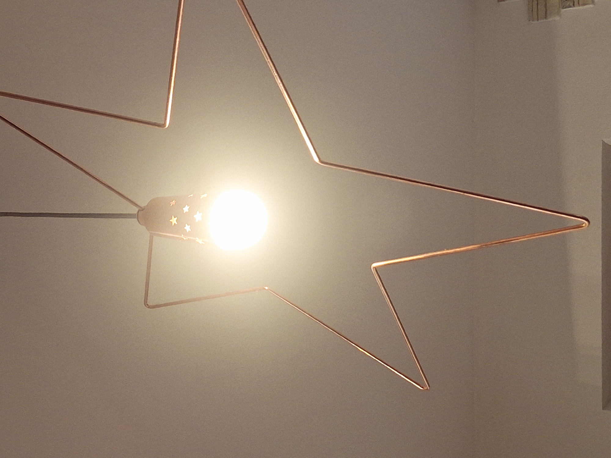 A low-angle, brightly lit interior shot features a star-shaped wireframe lamp with a lit bulb at its center. The lamp's wireframe is copper-colored, and the bulb fixture is decorated with small, star-shaped cutouts. The background is a plain, light-colored ceiling. No people are visible. The location of Chaikovsky, Russia, is not discernible from the image.
