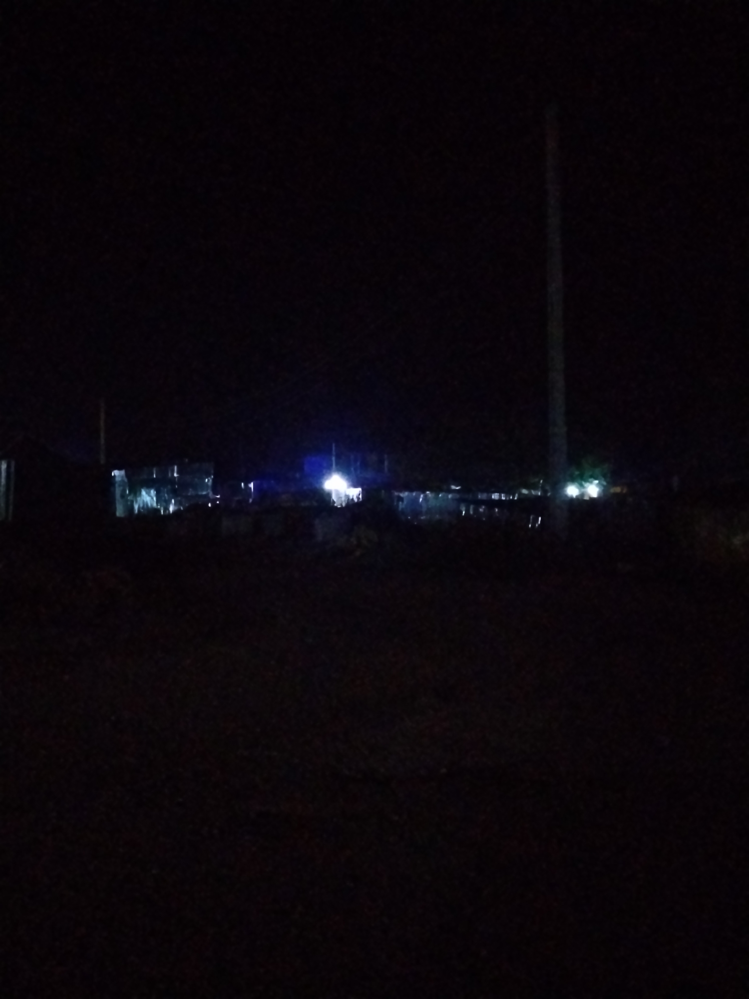 The image depicts a dimly lit outdoor scene at night, likely in the city of Durumi, Nigeria. The foreground is dark and unidentifiable, possibly rough terrain. In the midground, there appears to be a collection of structures, some with corrugated metal sheeting. Distant artificial lights are visible, including a bright blue light and several white lights, suggesting activity or illumination in the area. A tall, slender pole, possibly a utility pole, stands vertically on the right side of the image. The overall impression is that of a rural or semi-urban settlement at night, with minimal ambient light.