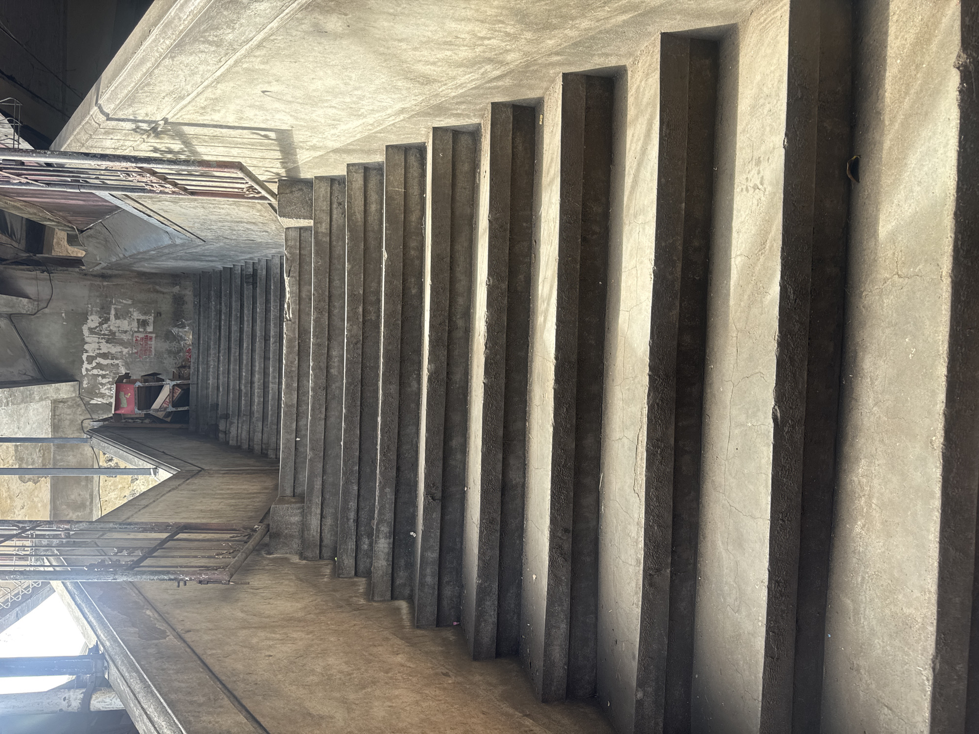 The image depicts a set of worn concrete stairs leading upwards in an urban environment. The stairs are wide and appear to be made of rough-textured concrete, with some cracks and wear visible on the surfaces. The lighting suggests it is daytime, with strong sunlight casting shadows on the steps and walls.
On the landing at the top of the stairs, to the right, there is a red metal cart with some stacked items, possibly refuse or stored goods. Further up, a person is partially visible sitting on a low structure, obscured by shadows and distance.
The walls flanking the stairs are also concrete, showing signs of age and weathering, with peeling paint or posters on the left wall. A metal railing, showing rust and discoloration, runs along the left side of the stairs, disappearing upwards. The overall impression is one of a somewhat neglected but functional public space.
This scene is located in Denpasar, Indonesia, as indicated by the context provided. Specifically, it appears to be a public staircase, possibly connecting different levels or areas within the city, such as a market, a pedestrian overpass, or a residential building access.