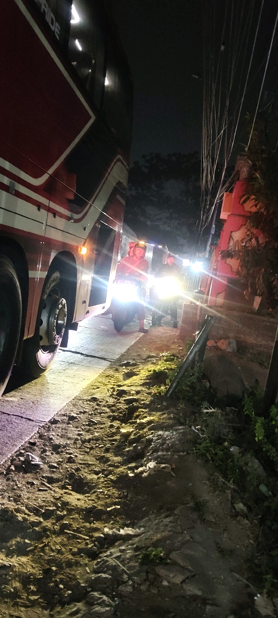 The image depicts a nighttime scene on a street in Sukaresmi, Indonesia. A large red and white bus is parked on the left side of the frame. In front of the bus, two motorcycles with bright headlights are visible. Several individuals are standing or crouching near the motorcycles, some of whom appear to be wearing helmets. The ground is unpaved and appears to be a dirt road with debris. Overhead, numerous power lines crisscross the sky. The lighting from the motorcycles creates lens flares and illuminates the immediate area. In the background, there are trees and a structure with red and orange elements.