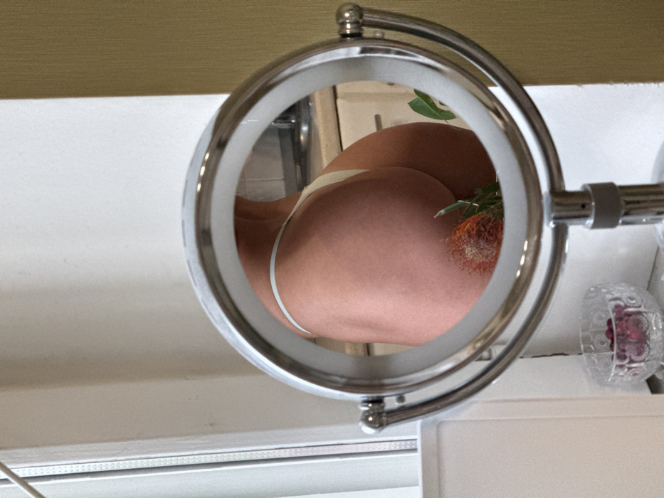 The image displays a chrome-finished, circular magnifying mirror with an illuminated outer rim, positioned on a white surface. Reflected in the mirror are the buttocks of an individual, appearing to be female, wearing light-colored, thong-style underwear. A protea flower, characterized by its reddish-orange, spiky bloom and green foliage, is positioned against the person's lower back/buttocks area. The background visible within the mirror's reflection suggests a room, possibly a kitchen or bathroom, with a metallic faucet and light-colored cabinets. To the left of the mirror, a white window frame is visible, and below it, a clear glass bowl containing red, spherical objects rests on the white surface. To the right of the mirror, a textured light brown wall is present. The scene is located indoors in Saint Petersburg, Russia.