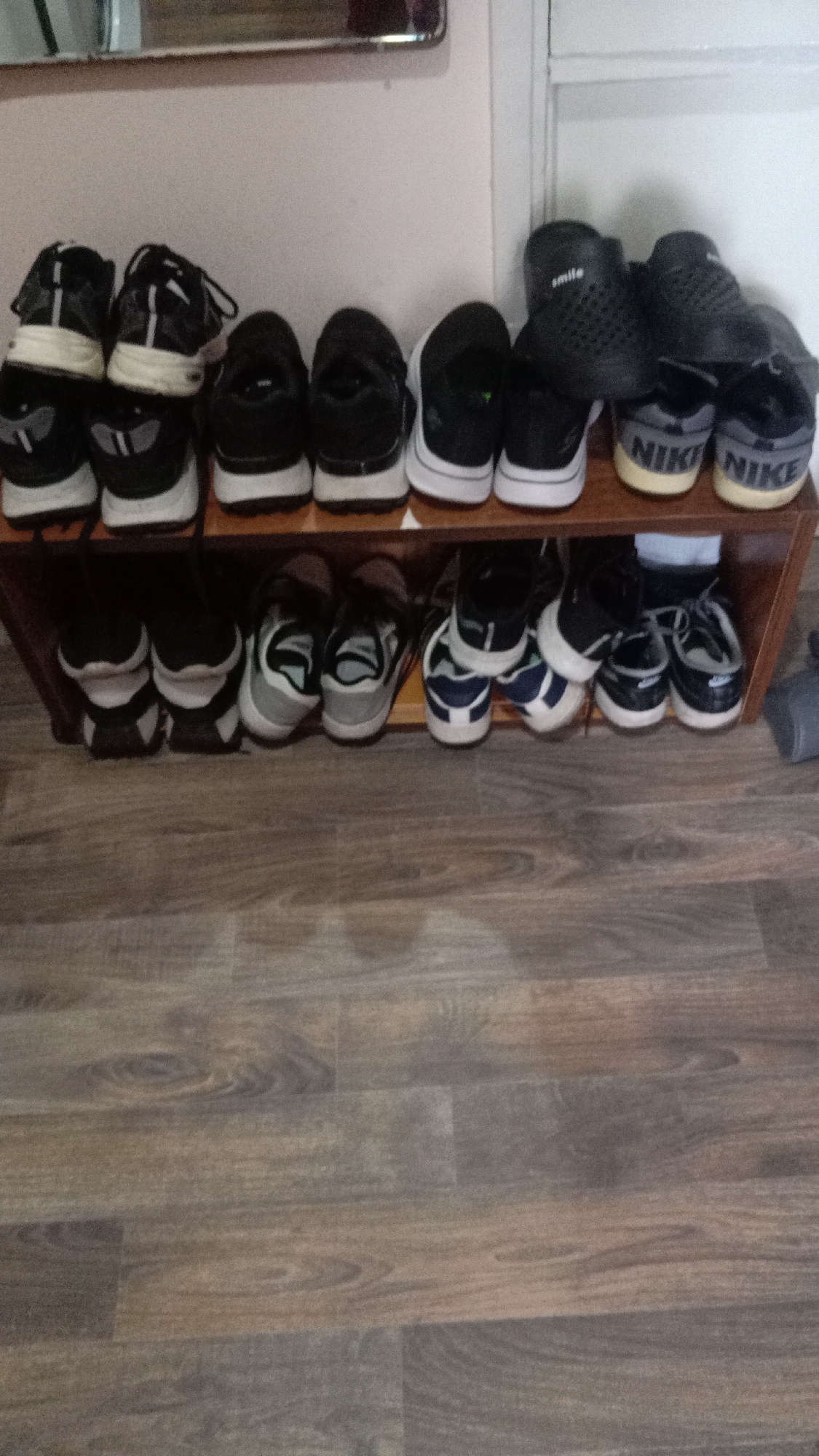 A medium shot depicts a brown, two-tiered shoe rack filled with various sneakers against a neutral-colored wall. The rack is positioned on a floor covered with brown wood-look planks. The top tier displays a collection of sneakers, including some black and white running shoes and a pair of black crocs with the word "smile" visible on one shoe. The bottom tier contains additional sneakers, predominantly gray and blue. A corner of what seems to be a mirror can be seen in the upper left corner of the frame. The photograph was likely taken indoors. The location of the image is Yerevan, Armenia.