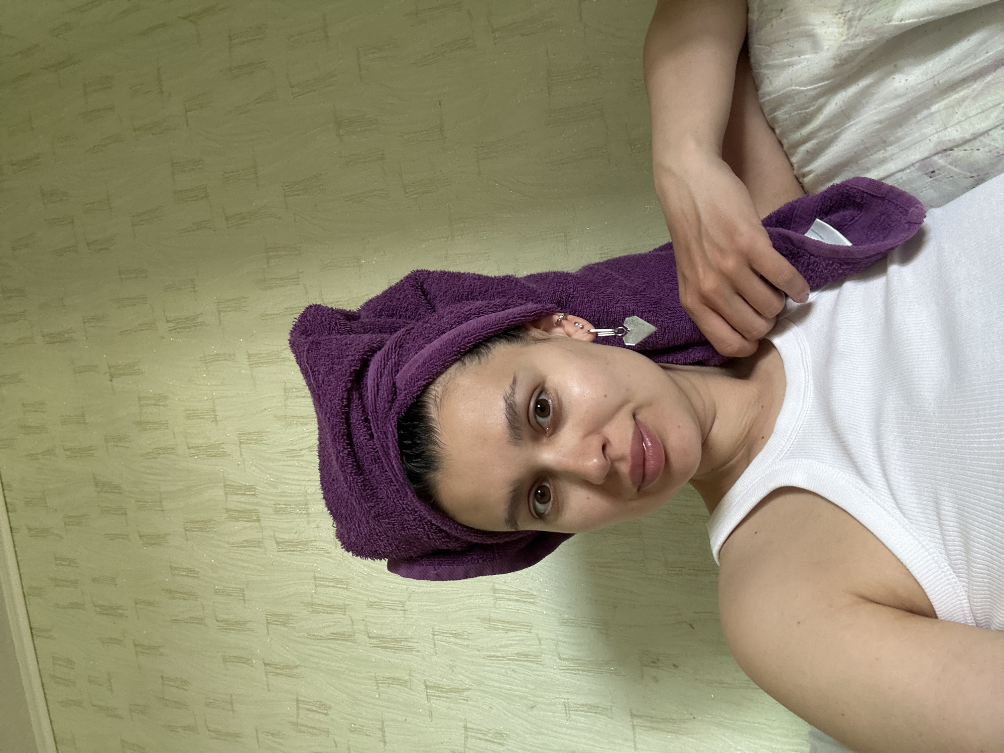 A woman is depicted indoors. She is wearing a white tank top and has a purple towel wrapped around her head. The background features a wall with a textured pattern. No details suggest the location is in the city of Chaikovsky, Russia. The image provides insufficient information to confirm the specific location.