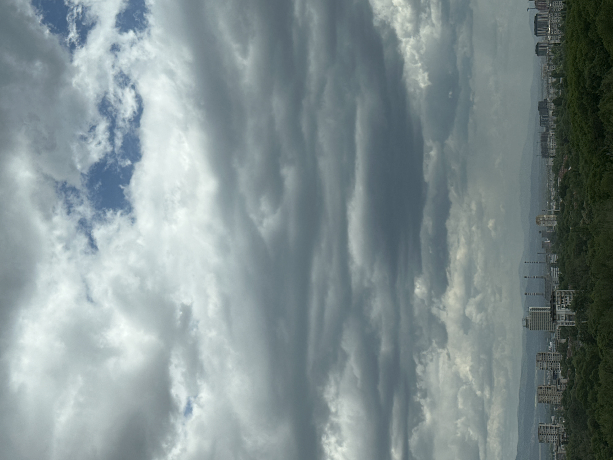 The image presents a vertically oriented landscape scene, featuring an overcast sky with dominant gray and white clouds. Interspersed within the clouds are glimpses of blue sky. In the lower portion of the frame, a city skyline is visible along the horizon, consisting of buildings and structures. Adjacent to the city, on the right side, lies a dense green area, likely a park or forest. The geographical location appears to be Sofia, Bulgaria, based on the architectural style of the buildings and the presence of tree-covered areas characteristic of the city.