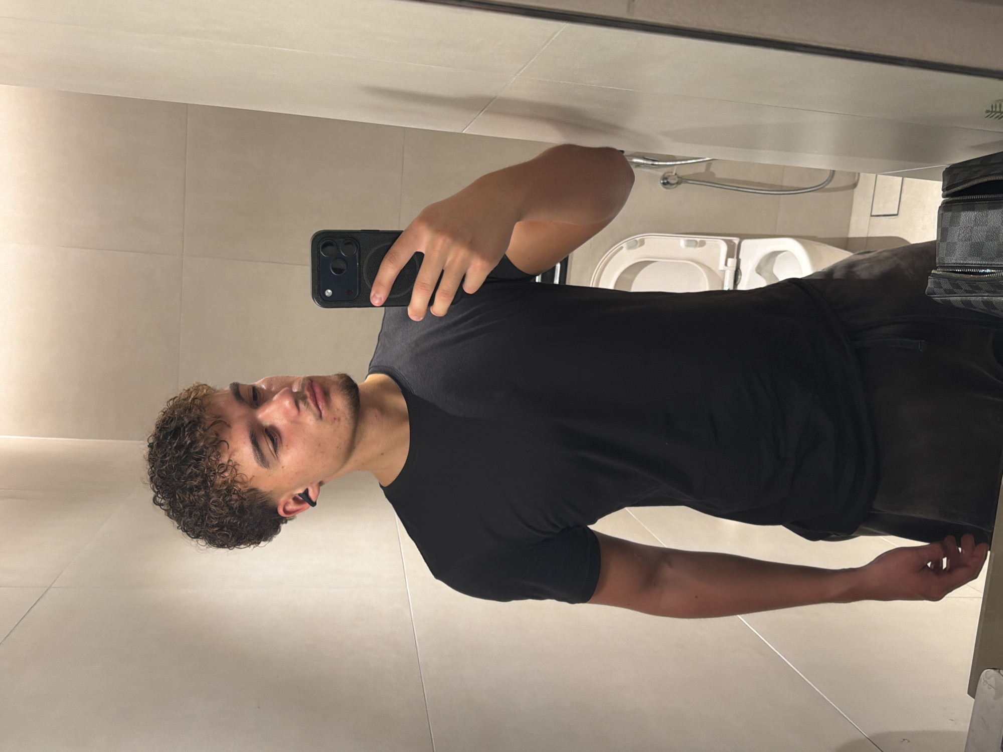 A young man is taking a selfie in a bathroom. He is wearing a black t-shirt and dark pants. He has curly hair. He is holding a black phone with multiple lenses. Behind him, a toilet and a shower are visible. A patterned bag is also visible. The scene is consistent with a bathroom interior. There's no information available that places this in Dubai, United Arab Emirates, so that part cannot be confirmed.