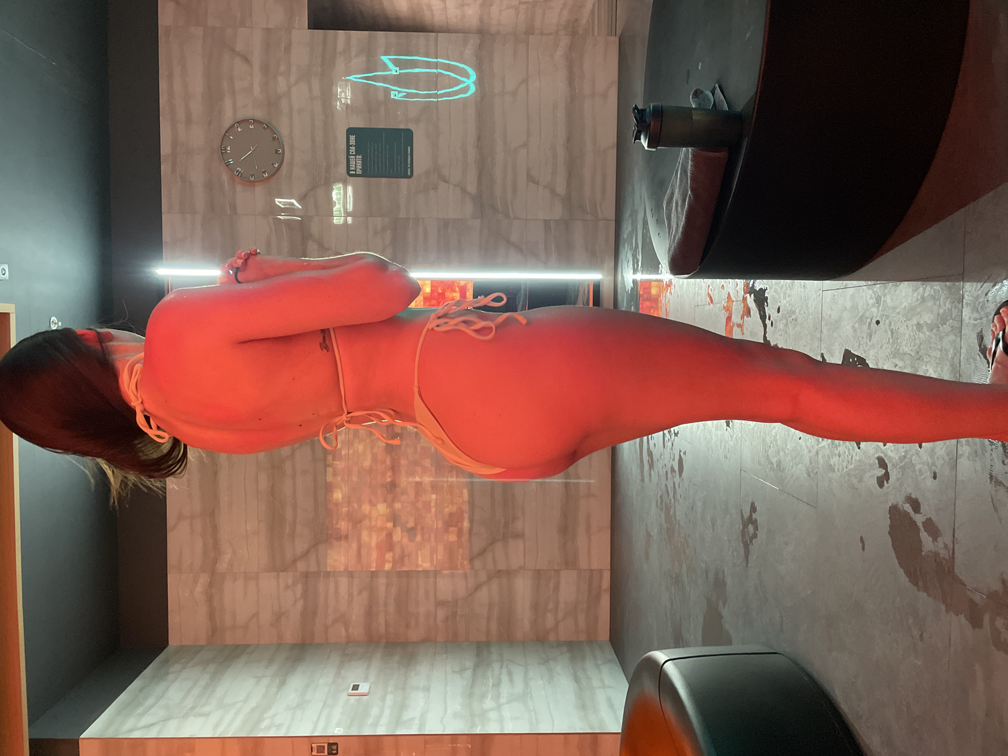 In a spa-like environment with marble-patterned walls and a dark ceiling, a woman wearing a yellow bikini stands facing away from the camera, illuminated by red lighting. She has dark hair and a tattoo on her left shoulder blade. The floor is tiled and wet, with visible water footprints. In the background, there is a round clock on the wall, a vertical neon light, and a sign in Russian with the text "В НАШЕЙ СПА-ЗОНЕ ПРИНЯТО" (In our spa zone it is customary). To the right, a black table holds a grey shaker bottle and a folded towel. To the left, a dark, curved object, possibly a seating area or a hot tub, is visible. The setting appears to be an indoor wellness or spa facility.
