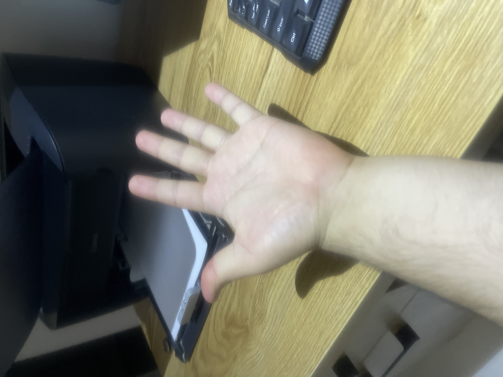 A human hand, palm facing the camera with fingers slightly splayed, is the primary subject in the foreground of this image. The hand appears to be pale and shows some faint red or pinkish discoloration, possibly from pressure or irritation. The arm is visible extending downwards, showing light hair growth. The scene is set on a wooden desk. In the background, a black Canon printer is visible with a piece of paper partially inserted into its output tray. To the right of the hand, a portion of a black keyboard with visible keys like "Esc", "Tab", "Caps Lock", "Shift", and "Ctrl" is present. The environment suggests an indoor setting, likely an office or study area. The image was taken in Qarshi, Uzbekistan.