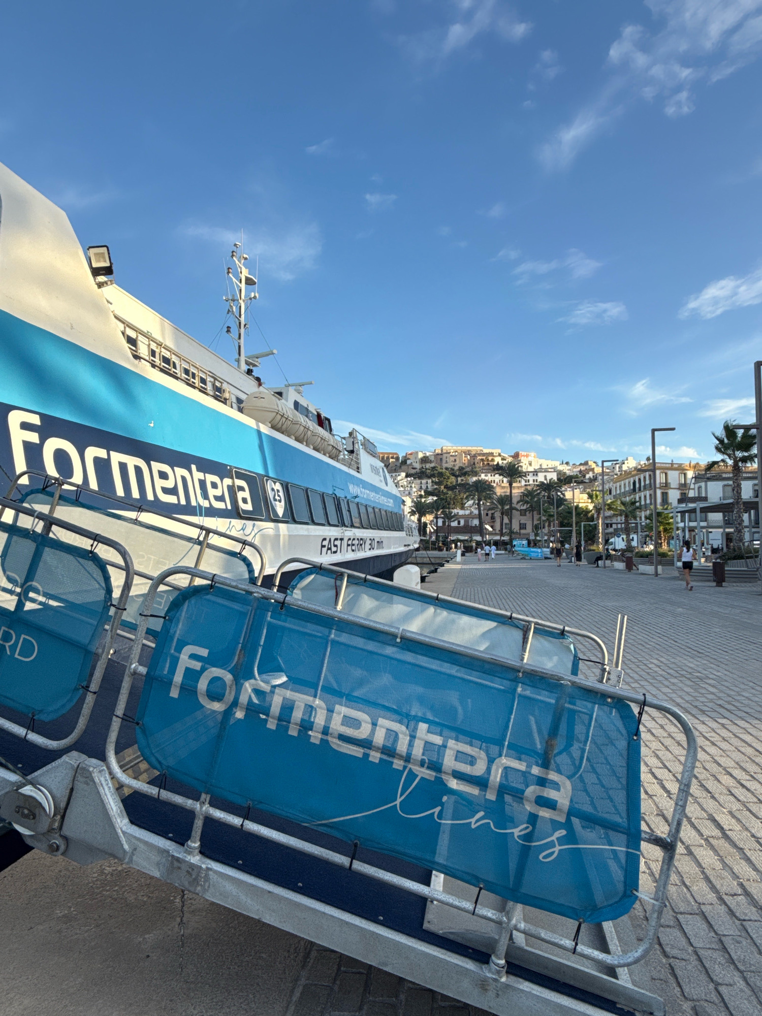A wide-angle, low-angle shot captures a blue and white fast ferry docked at a harbor in Ibiza, Spain, under a clear blue sky with scattered clouds. The ferry, named "Formentera Lines," dominates the left side of the frame, with its name prominently displayed in large white lettering. In the foreground, a metal ramp with blue mesh barriers, also bearing the "Formentera Lines" logo, leads towards the ferry.
Beyond the ferry, a paved promenade lined with palm trees stretches into the distance. A low wall separates the promenade from the water. Buildings with white and ochre facades, characteristic of Ibiza's architecture, are visible on a hillside in the background, suggesting a town or city setting. Several people are seen walking on the promenade, and a woman in a dark top and shorts walks away from the camera. The scene is well-lit, indicating it is daytime. The presence of the fast ferry and the urban backdrop strongly suggest a port or harbor area in the city of Ibiza, likely near the ferry terminal.