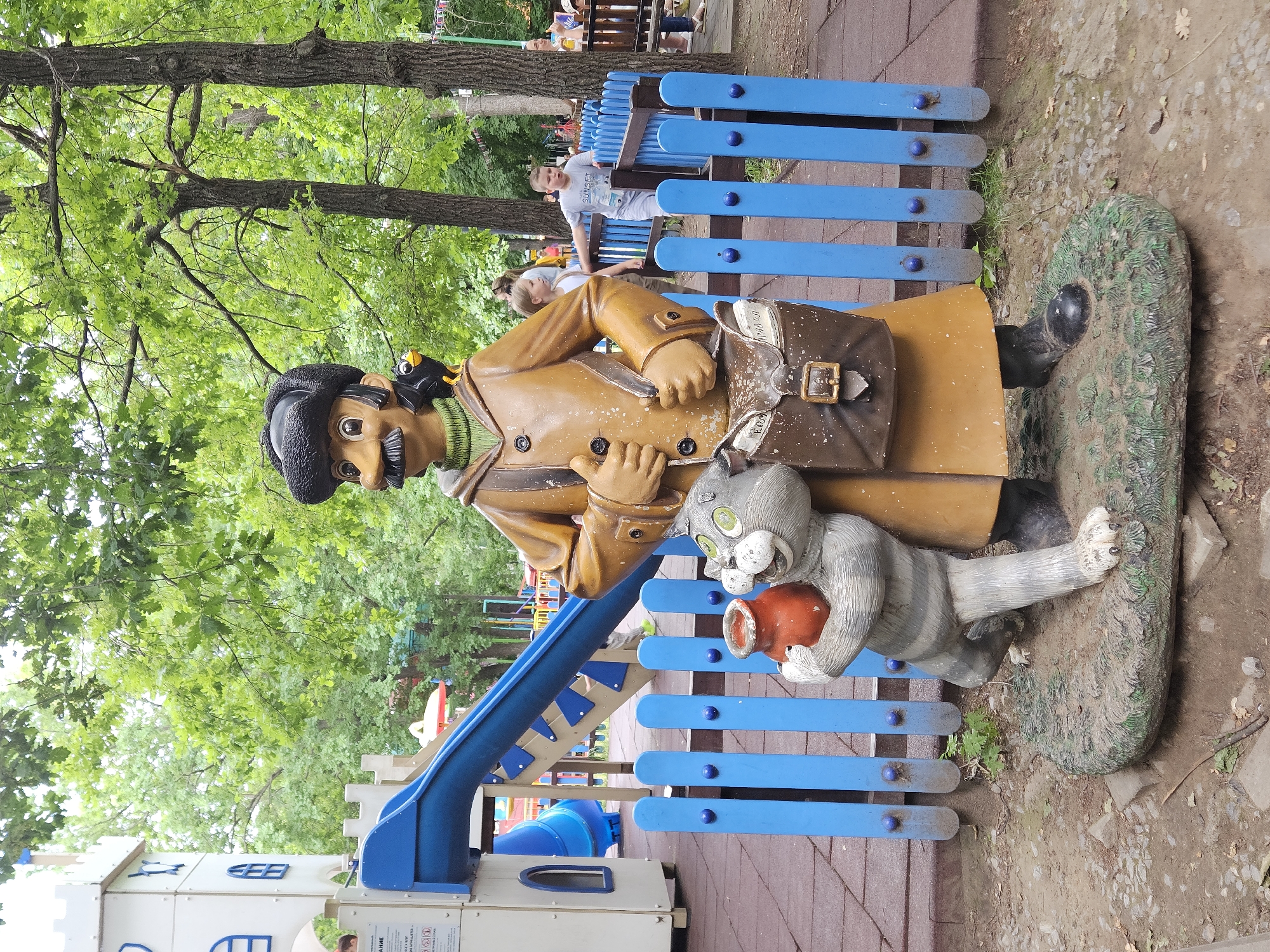 A statue of a character from a children's story, possibly "The Cat and the Rat" or "The House That Jack Built," stands in a park in Ulyanovsk, Russia. The statue depicts a man in a brown coat and a black hat, holding a cat that is clutching a pot. A small black bird with a yellow beak perches on the man's shoulder. Behind the statue is a blue fence and a playground with slides. The ground is covered with dirt and scattered leaves. In the background, trees with green foliage form a canopy over the park. Several children are visible in the background, playing on the playground. The atmosphere is bright and sunny, suggesting it is a warm day.