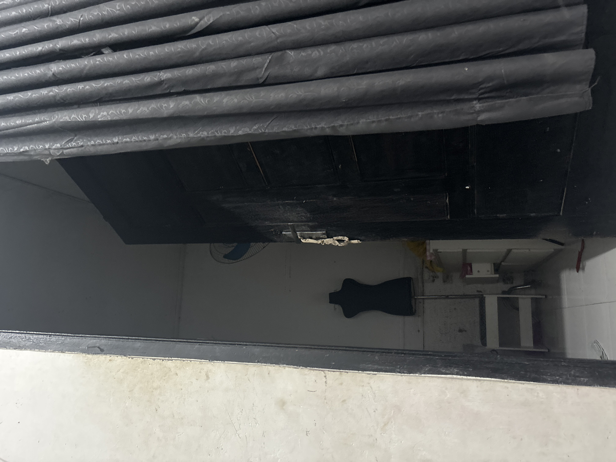 The image shows an interior view looking through an open doorway into a room. The room is dimly lit, with a black mannequin visible in the center. To the right of the doorway, a dark curtain hangs, obscuring part of the view. A fan is visible on the back wall of the room. The overall scene suggests a storage or workshop area, possibly in a residential building in North Jakarta, Indonesia. The walls appear to be painted white, and the floor is tiled. There is a shelf unit visible in the background with some items on it.