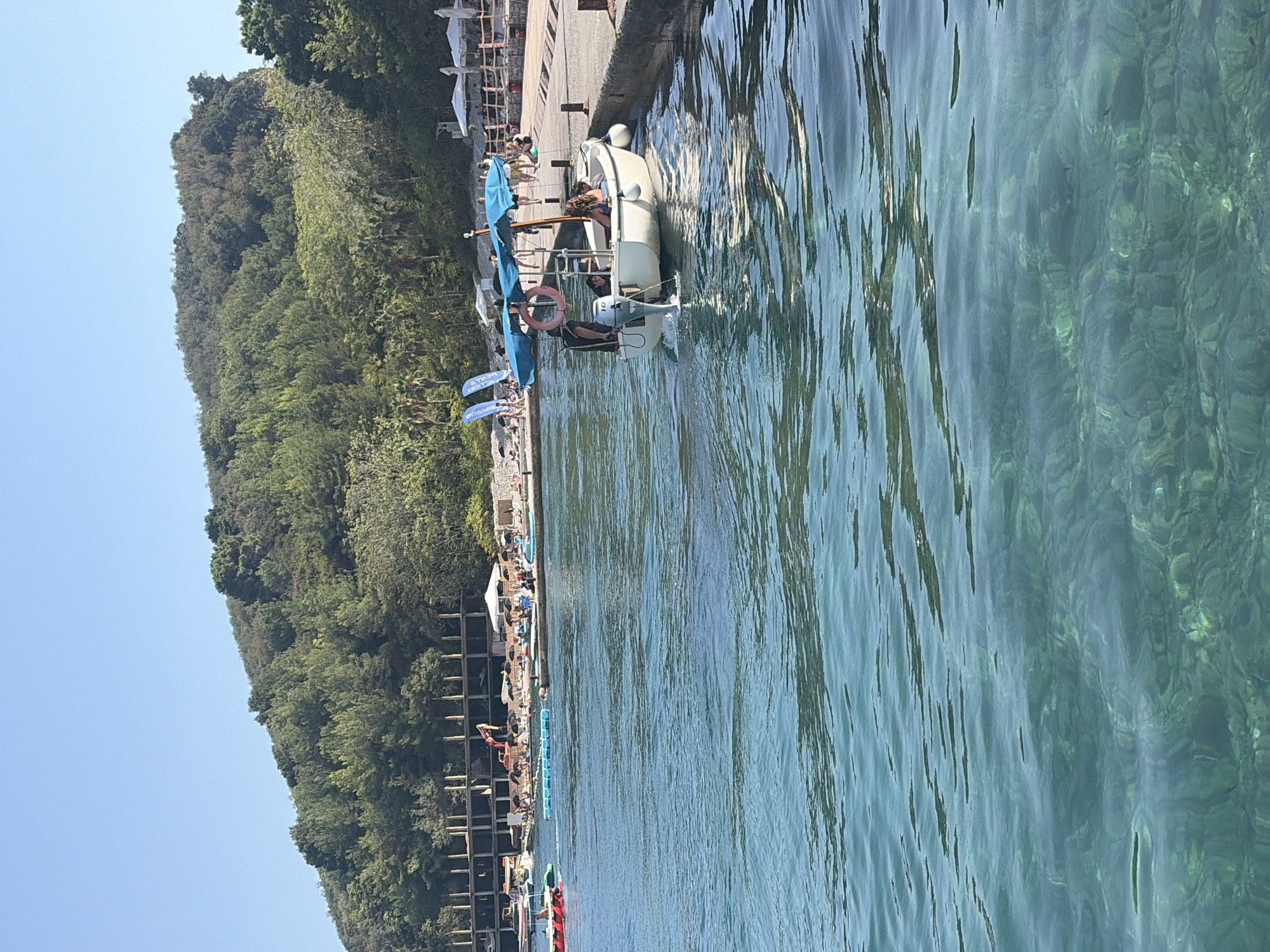 The image depicts a bright, sunny day at a coastal location, likely Budva, Montenegro, given the distinct hillside covered in lush green trees that slopes down towards the sea. The water is clear and turquoise, revealing the rocky seabed beneath.
In the foreground, the water is rippling, reflecting the sunlight. Closer to the shore, a white motorboat with a blue canopy is docked or slowly moving along a concrete pier. Three people are visible on the boat; one person is at the stern, operating the outboard motor, and two passengers are seated in the boat, one facing forward and the other looking towards the camera. The boat has a life preserver ring attached to its canopy.
Along the shore, there is a paved walkway or pier with wooden railings in some sections. Beyond the walkway, a beach area is visible with numerous beach umbrellas and people relaxing. Some beach chairs are set up. In the background, a multi-story building with an exposed concrete frame appears to be under construction or renovation. A red construction vehicle, possibly an excavator, is visible near this building. Further back, the dense, green forest covers the hillside, with the clear blue sky above. Several other small boats and floating water toys are present in the water near the beach.