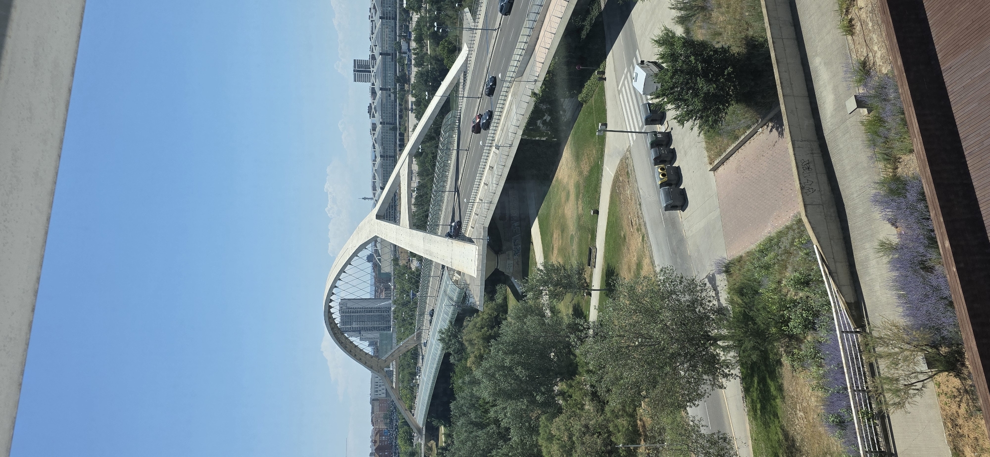 An aerial, wide shot captures a modern, concrete bridge with an arched main span over a clear blue sky in Zaragoza, Spain. The bridge, which has a unique, sweeping design, supports multiple lanes of traffic, with several cars visible driving on the roadway. The bridge extends across a verdant landscape of trees and green spaces. In the background, the cityscape of Zaragoza is visible, featuring buildings and what appears to be a stadium or large event venue. The foreground shows a paved area with decorative purple flowers and vegetation, along with several large, dark-colored trash receptacles. The scene is bathed in bright sunlight, indicating a clear day.