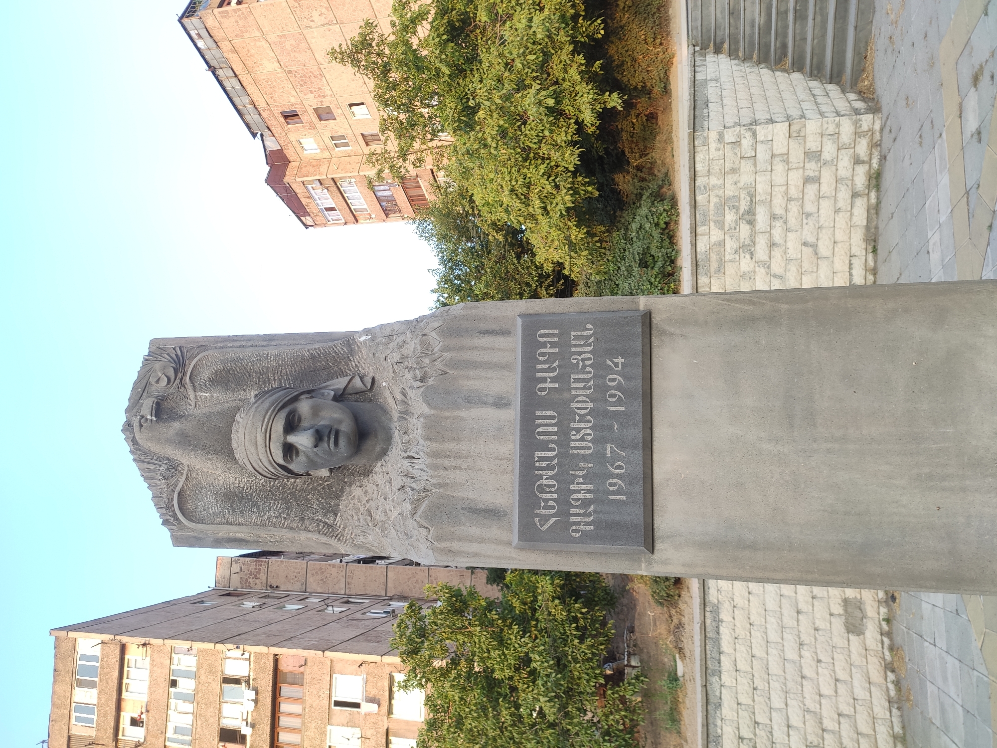 This is a front-facing, eye-level, medium shot of a monument in Yerevan, Armenia. The monument, made of dark gray stone, features a carved bust of a man with a headscarf, positioned behind a plaque with Armenian script and dates. The man's head faces forward, and the overall style suggests a memorial or artistic representation. Behind the monument, two apartment buildings with concrete facades and numerous windows are visible. The buildings are residential and appear somewhat aged. Trees with green foliage are situated between the monument and the buildings. The sky is clear and light blue, indicating daytime. The ground in front of the monument is paved with square tiles, and there is a stone wall with stairs leading up to the right of the frame. The monument is likely located in a public space, possibly a park or square, given the presence of paved walkways and stairs.