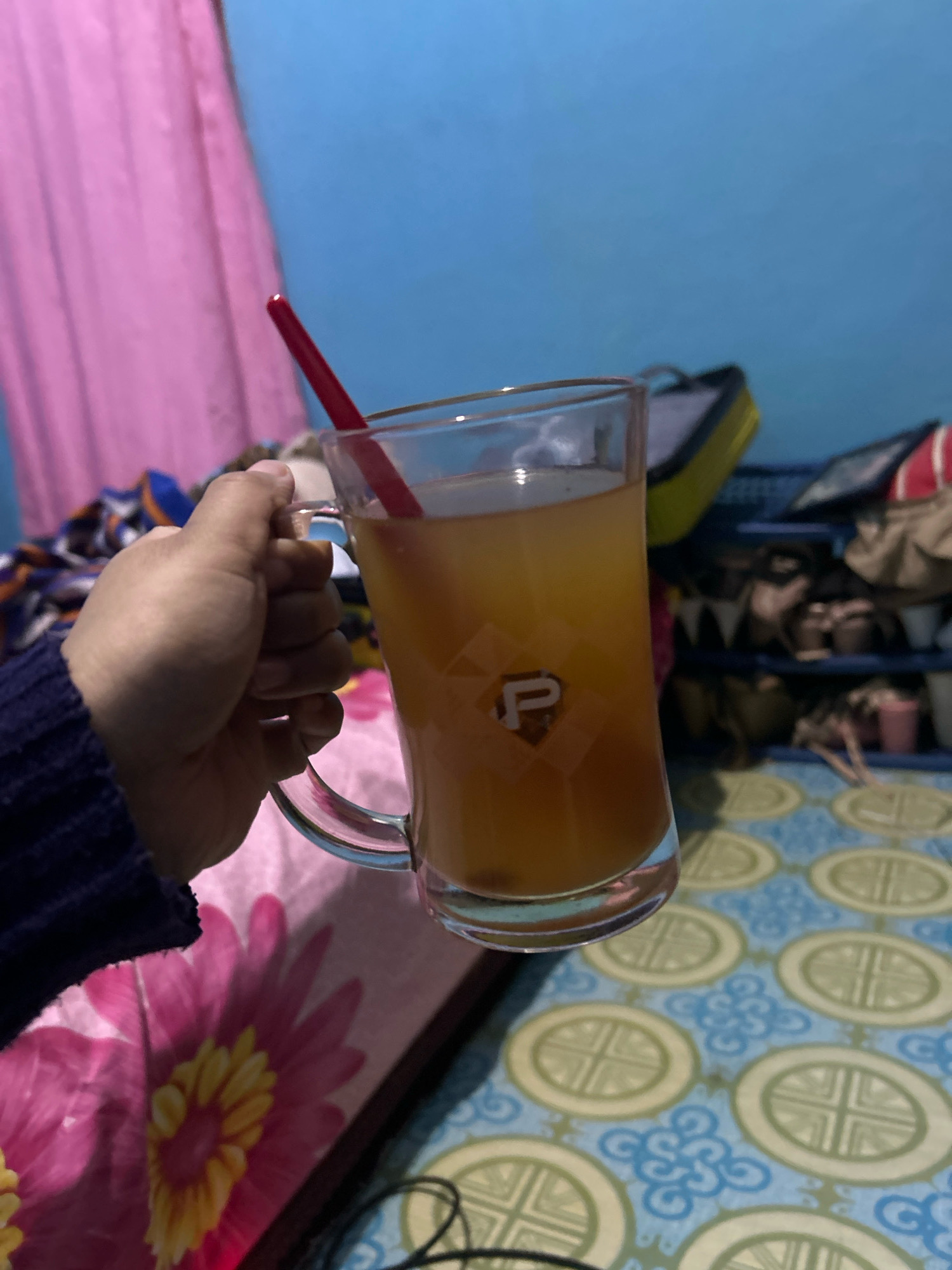 In Cihaurbeuti, Indonesia, a hand wearing a dark blue sweater holds a glass mug filled with amber-colored liquid and a red straw. The mug has a diamond-shaped emblem with the letter "P" inside. In the background, a pink curtain hangs to the left, and a blue wall is visible above. To the right, a shelf unit holds various items, including shoes and bags. The surface beneath the mug is covered with a blue and yellow patterned fabric.