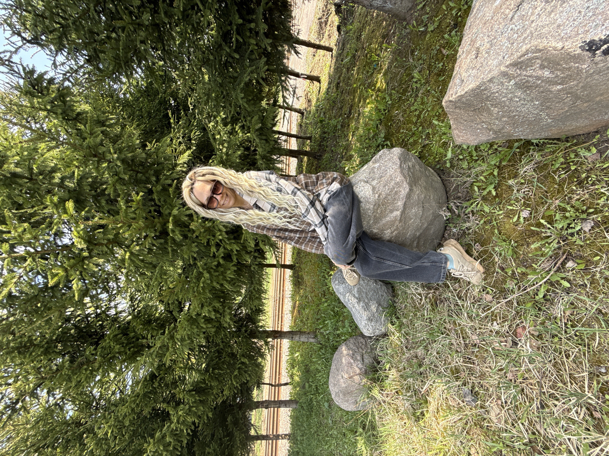 A person with long blonde hair, wearing sunglasses, a plaid shirt, and jeans, is reclining on a large gray rock amidst a grassy area with several other rocks. A line of evergreen trees is visible to the left of the frame, with glimpses of railroad tracks behind them. The location is Rusanovo, Russia.