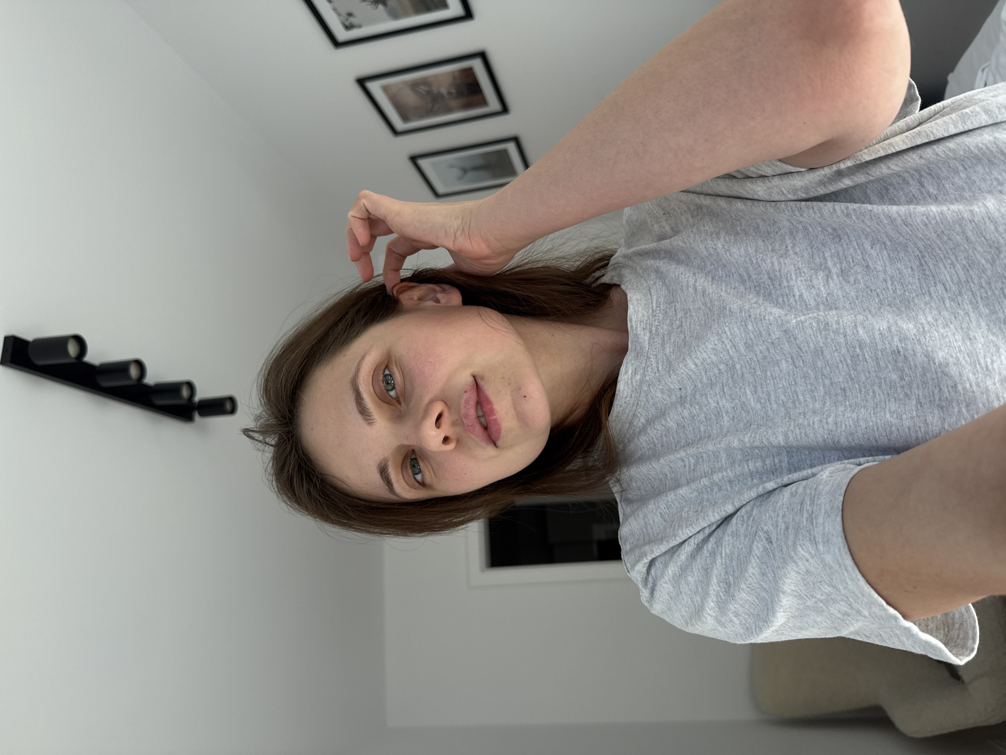 The media file shows a young woman indoors, with her head tilted and her right hand touching her hair near her ear. She is looking towards the camera with a soft expression. She is wearing a light grey t-shirt. In the background, on a white wall, there are three framed pictures hung vertically. To the left of the woman, a black wall-mounted object with four cylindrical elements is visible. The scene appears to be a room, possibly a bedroom or living room, with neutral-colored walls. The lighting suggests it is daytime. The overall mood is casual and relaxed. No visible text is present.